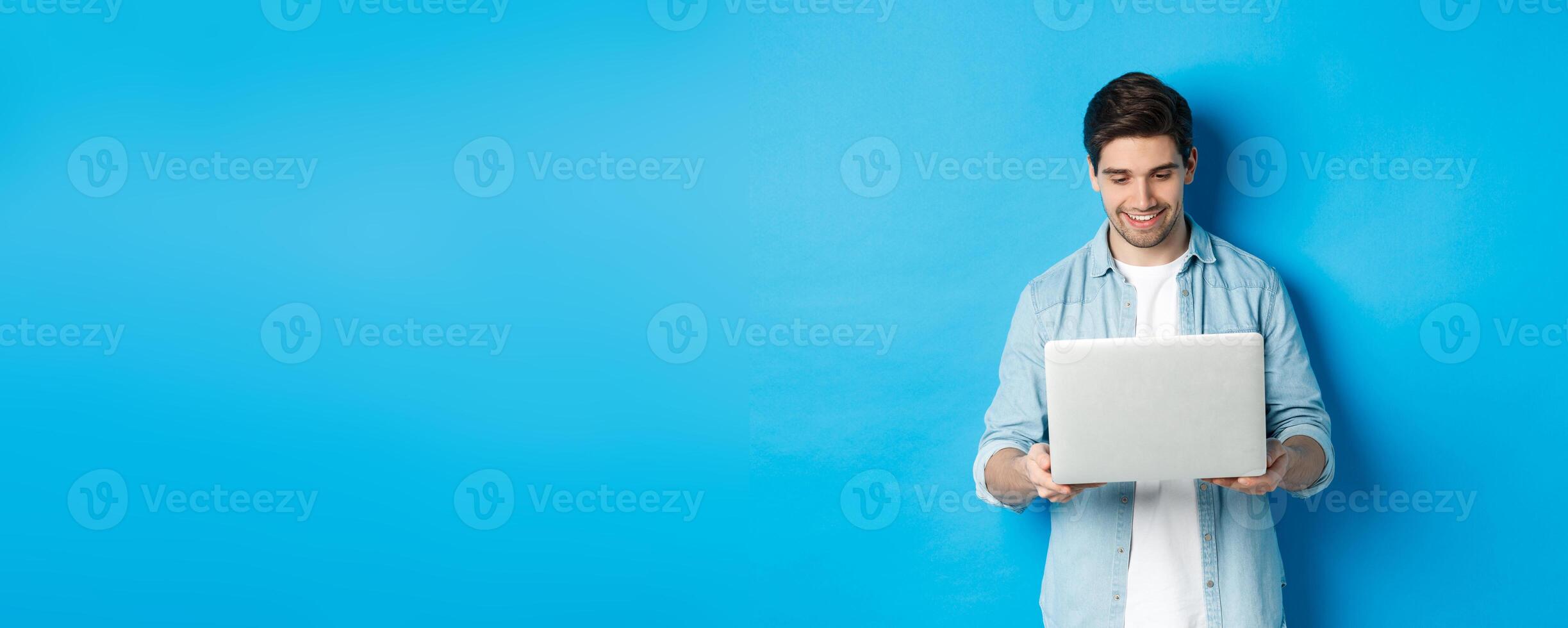 Handsome man working on laptop, smiling and looking at screen satisfied, standing against blue background photo