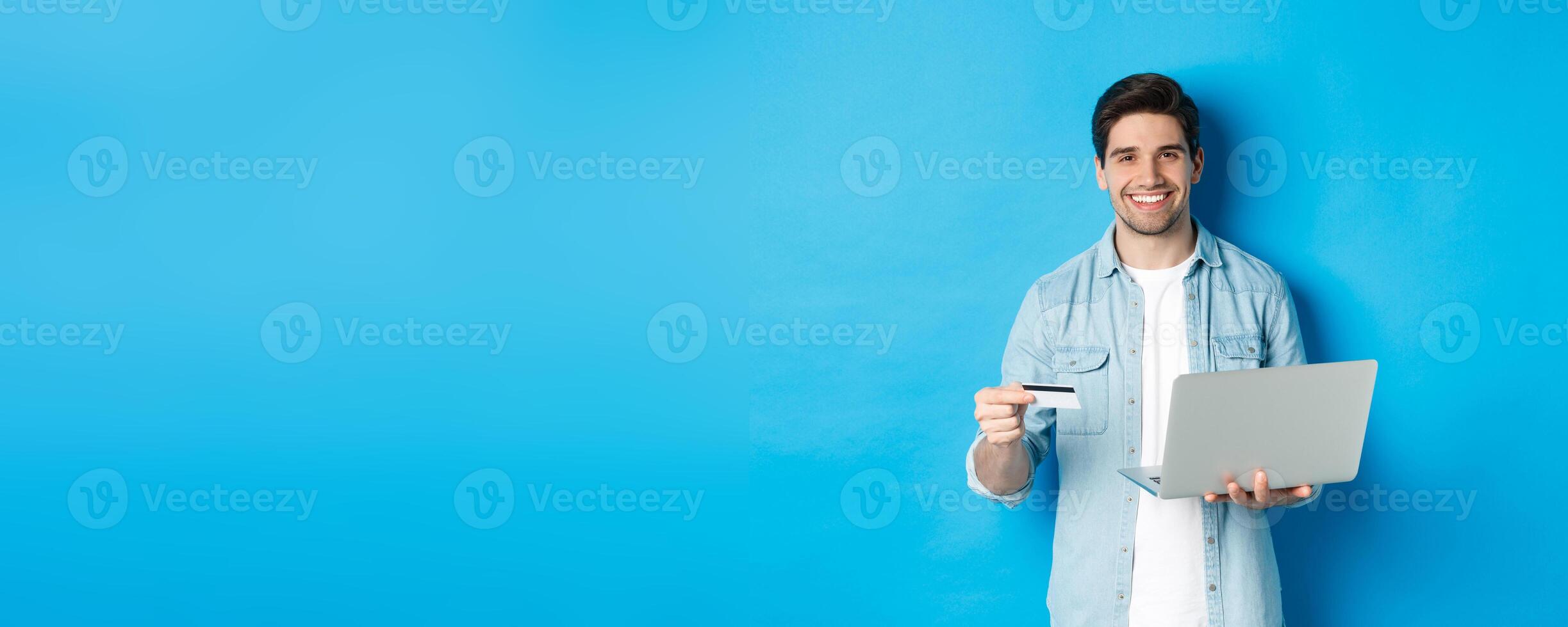 Handsome man smiling, shopping online and paying for product, holding credit card with laptop, standing over blue background photo