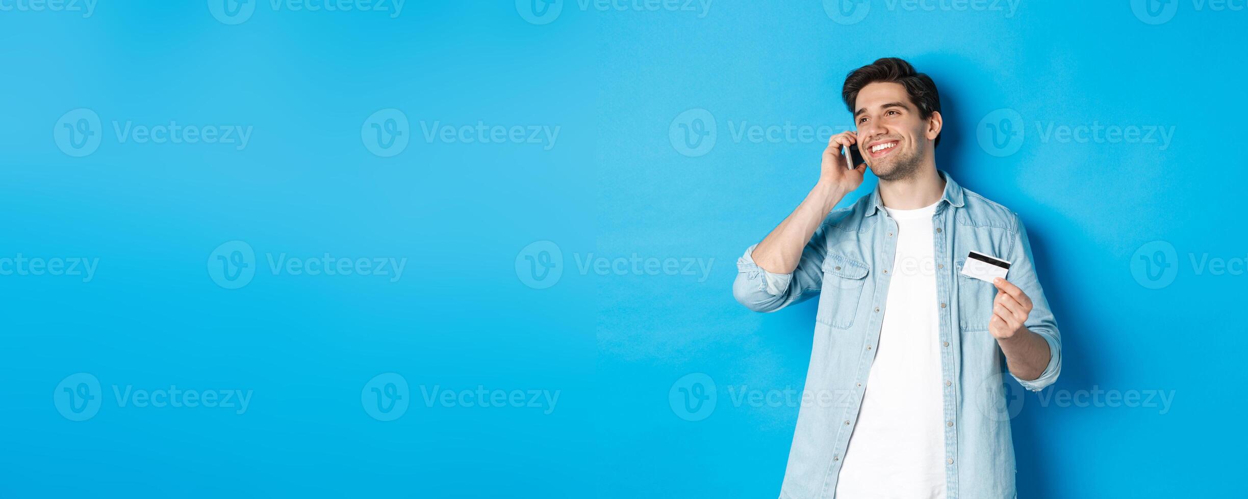 Smiling man call bank support, talking on mobile phone and holding credit card, standing against blue background photo