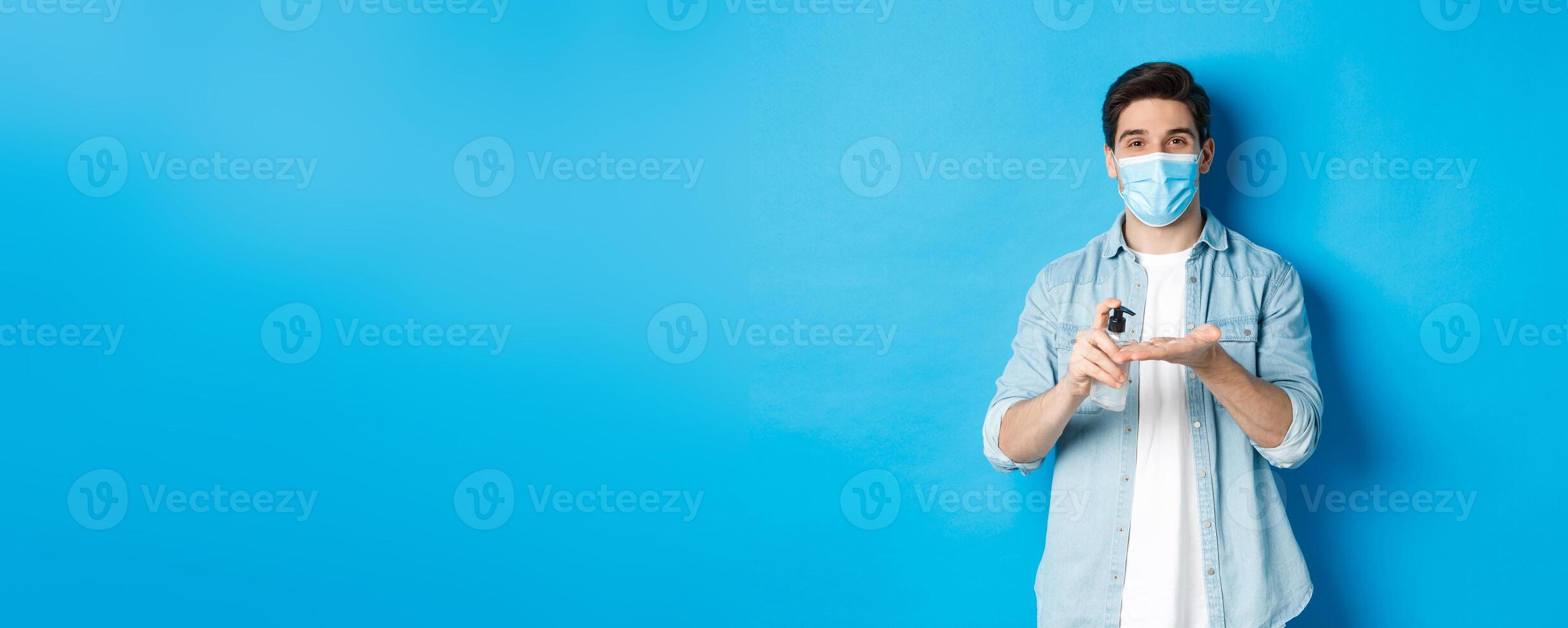 Concept of covid-19, pandemic and social distancing. Handsome young man in medical mask disinfecting hands with sanitizer, using antiseptic, standing against blue background photo