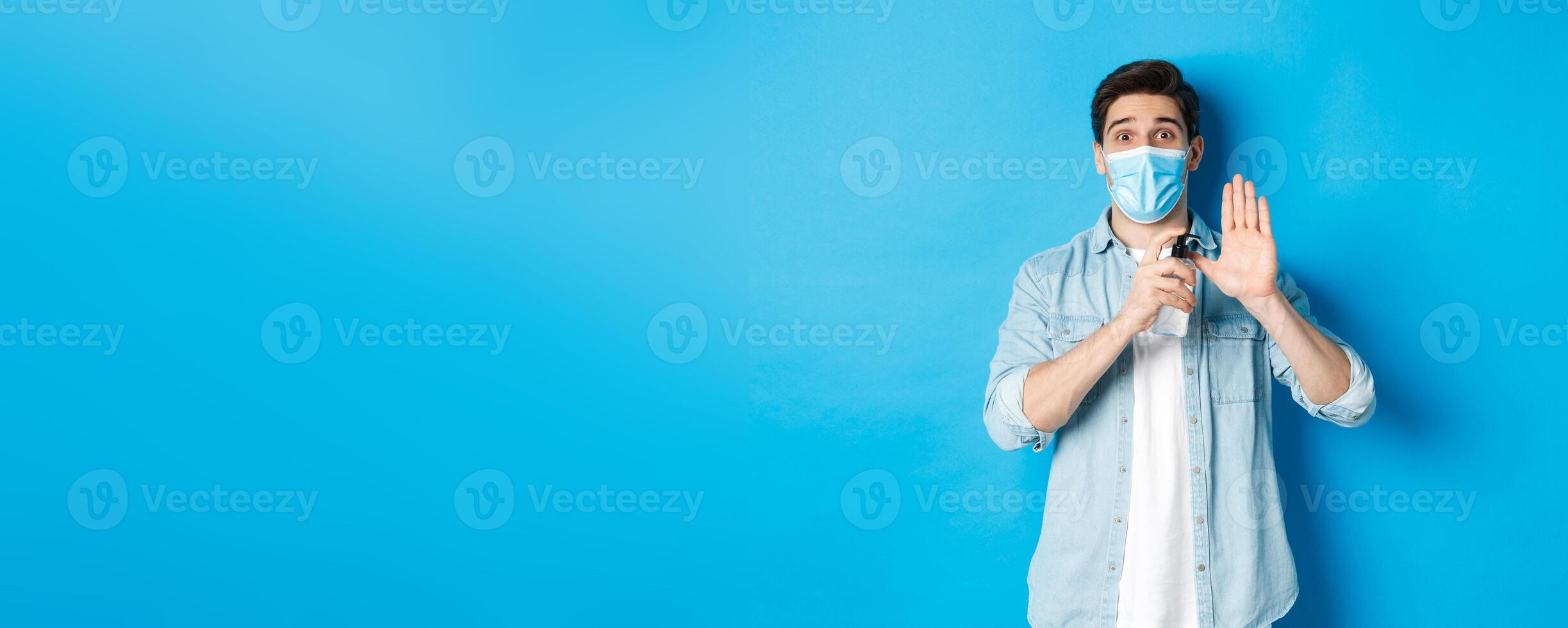 Concept of covid-19, pandemic and social distancing. Cheerful guy in medical mask showing how to disinfect hands with sanitizer, using antiseptic, preventing virus spread, blue background photo