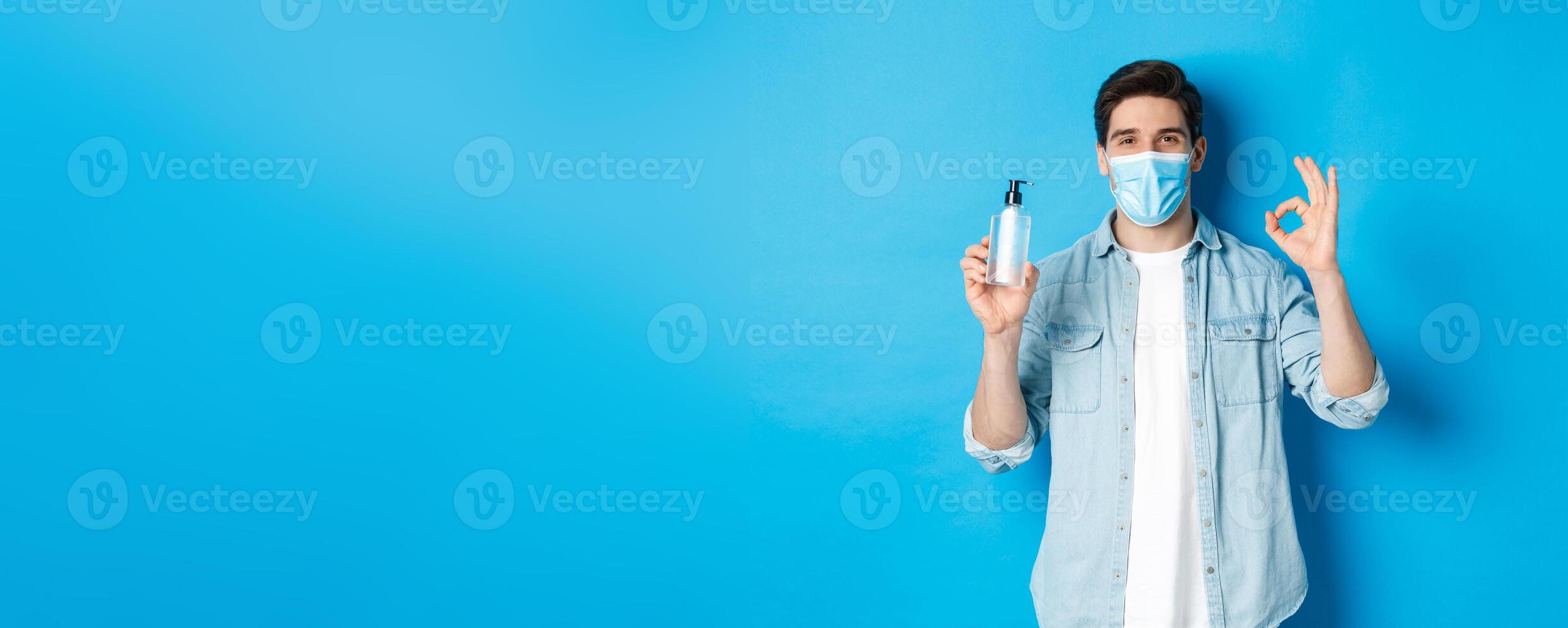 Concept of covid-19, pandemic and social distancing. Satisfied young man in medical mask recommending hand sanitizer, showing ok sign and antiseptic, standing against blue background photo
