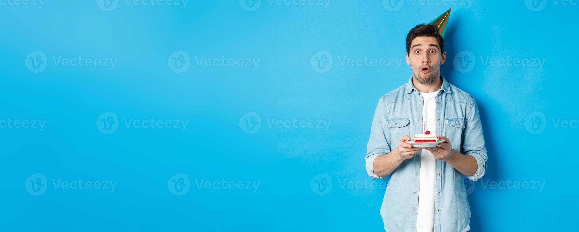 Handsome man in party cone holding birthday cake, looking surprised, standing over blue background photo