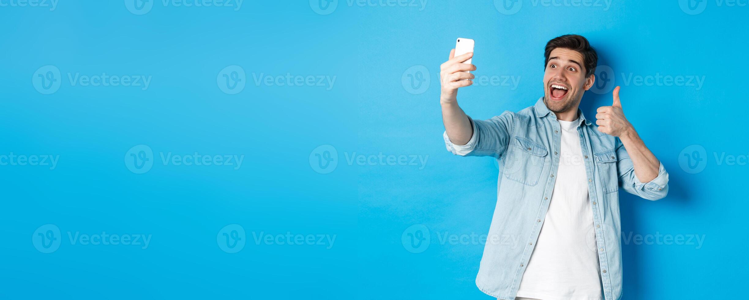 Happy man taking selfie and showing thumb up in approval on blue background, holding mobile phone photo