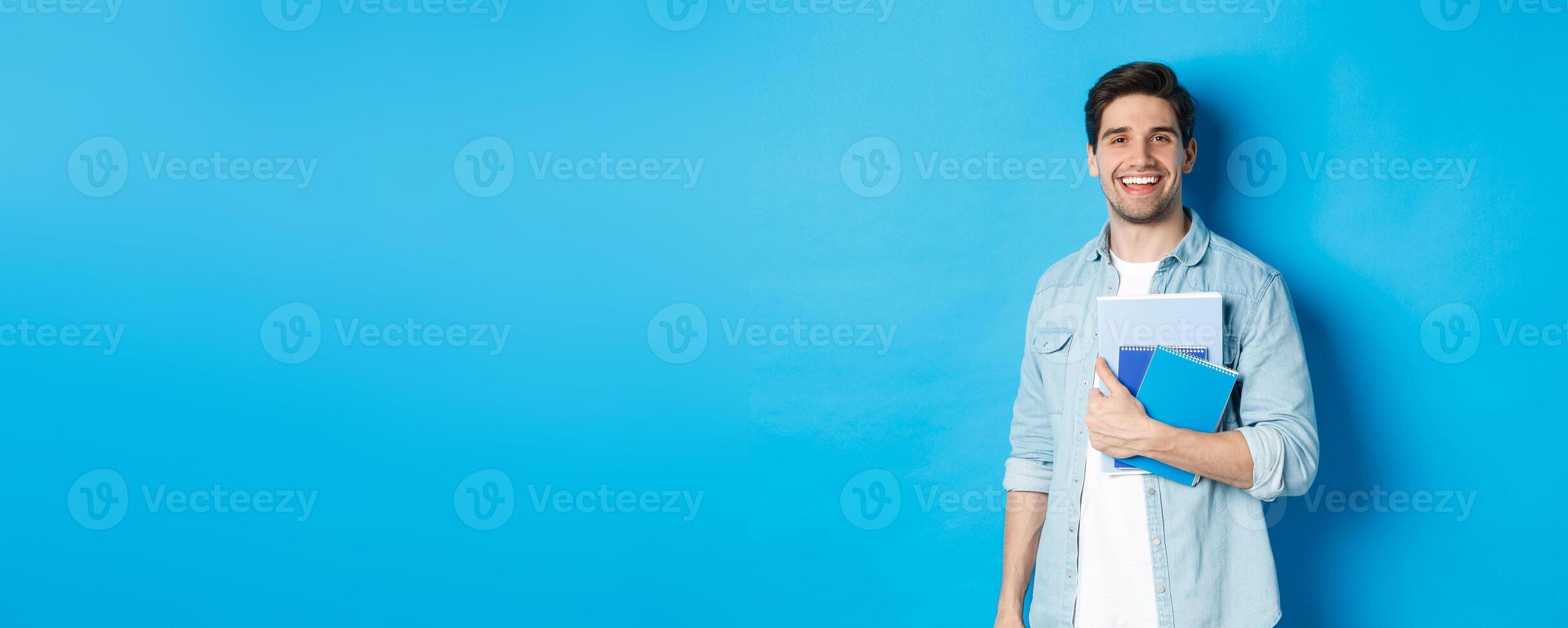 Smiling man studying, holding notebooks and looking happy, standing over blue background photo