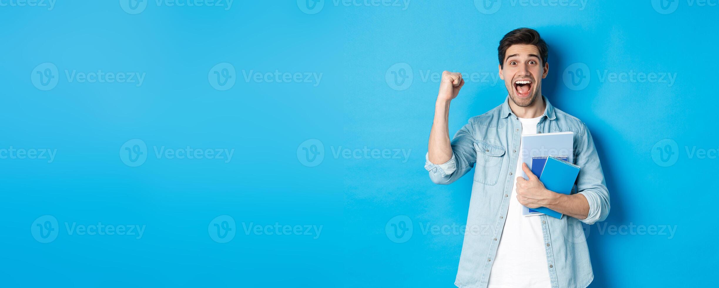 Cheerful guy holding notebooks and celebrating, making fist pump and shouting yes with excitement, standing over blue background photo