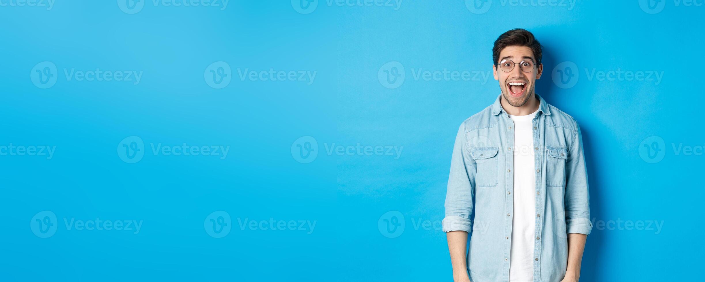 Attractive happy man in glasses looking surprised, checking out advertisement, standing over blue background photo
