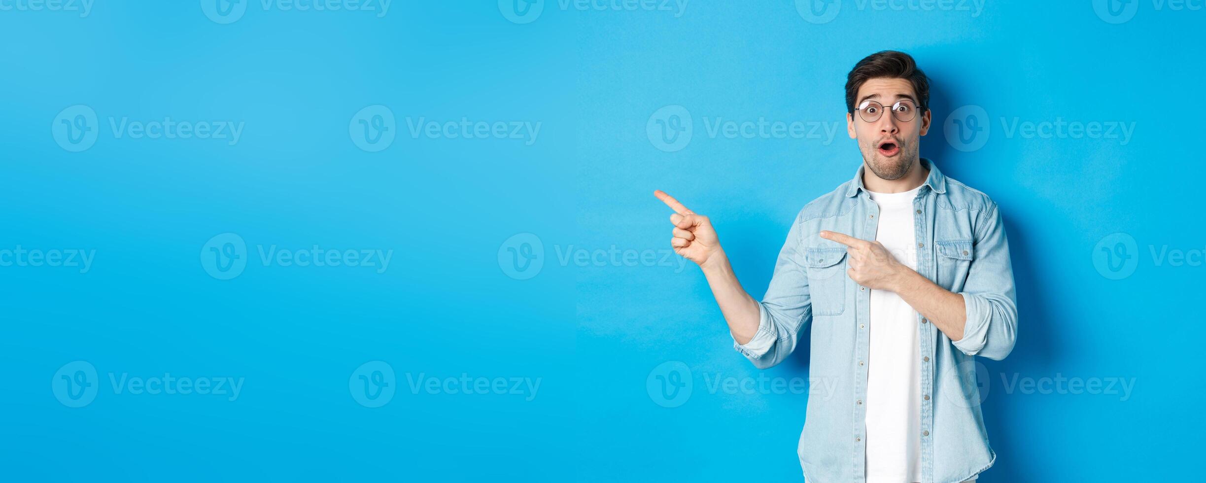 Surprised man in glasses pointing fingers left at copy space, asking question about product, looking intrigued, standing over blue background photo