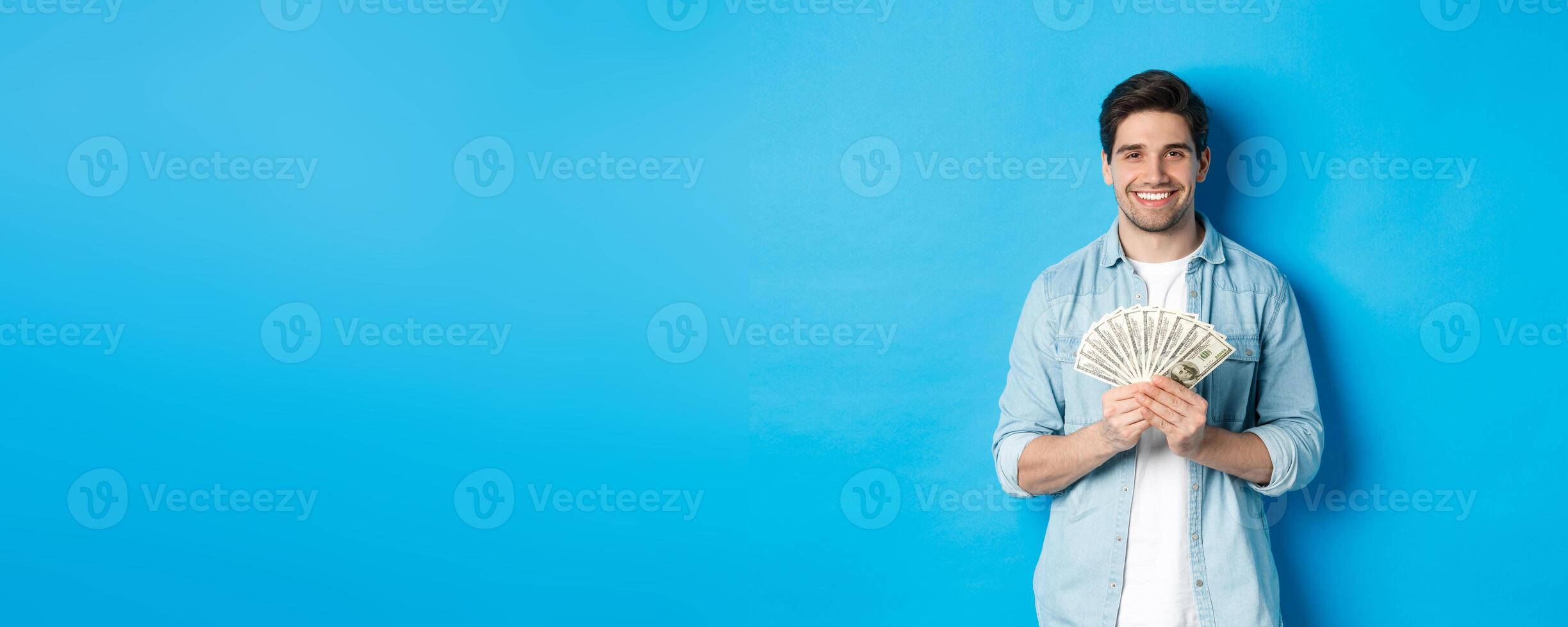Handsome smiling man holding money, concept of finance and banking, standing over blue background photo