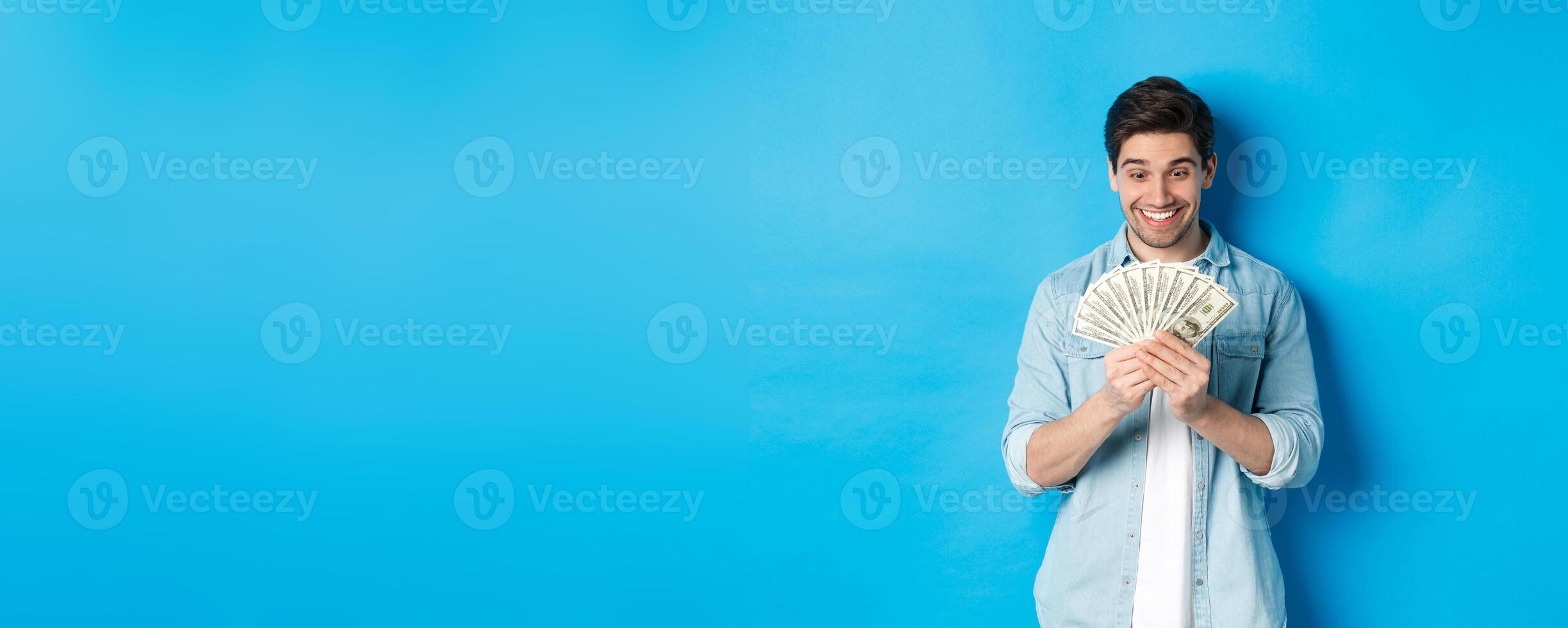 Excited successful man counting money, looking satisfied at cash and smiling, standing over blue background photo
