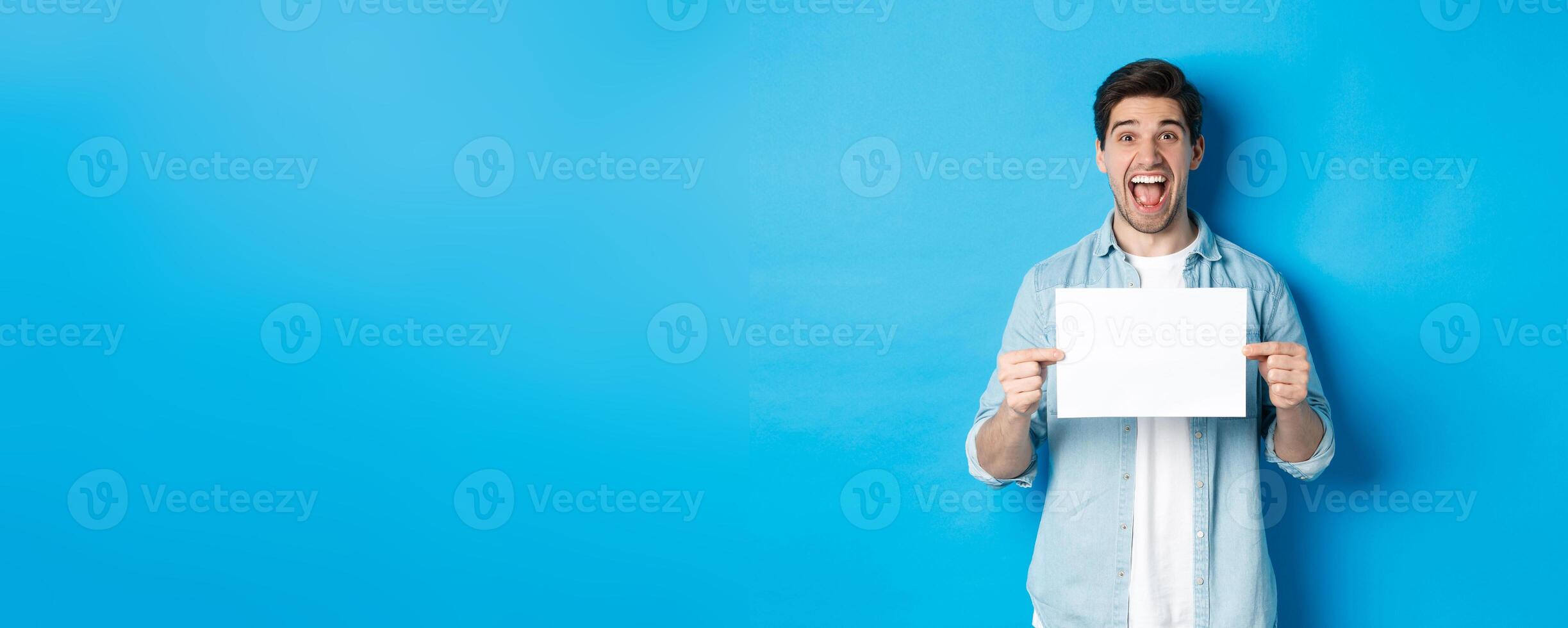 Happy attractive man showing piece of paper for your logo sign, standing amazed against blue background photo