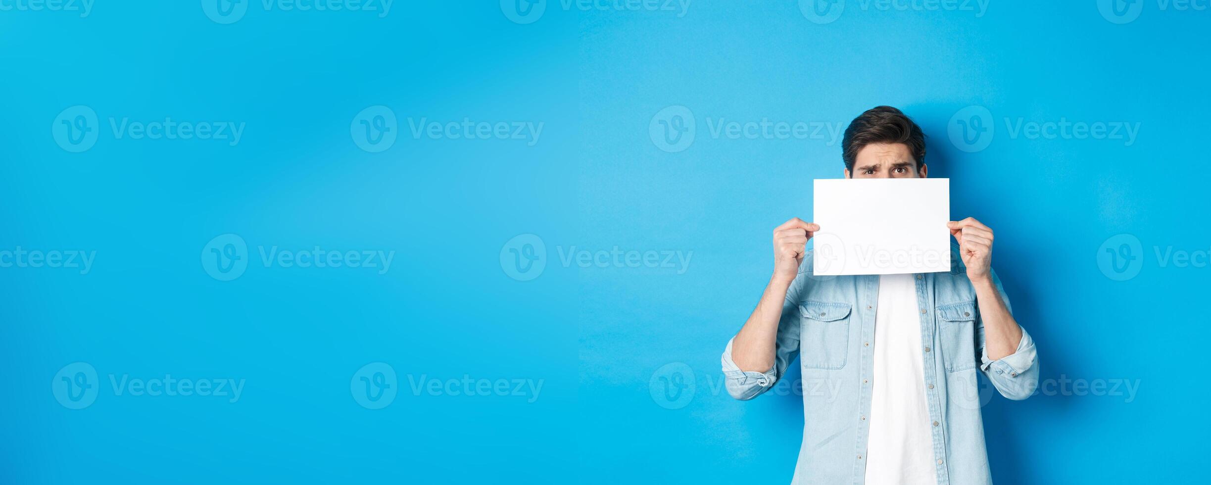 Shy and nervous guy hiding face behind blank piece of paper, peeking with worried look, showing your logo, standing over blue background photo