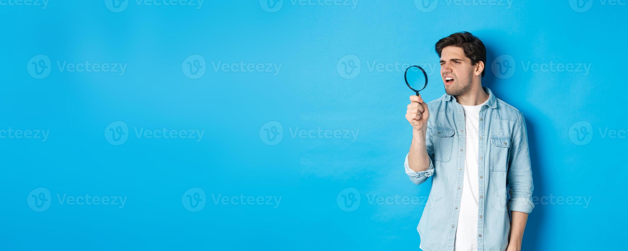 Young thoughtful guy looking through magnifying glass, reading something tiny, standing over blue background photo