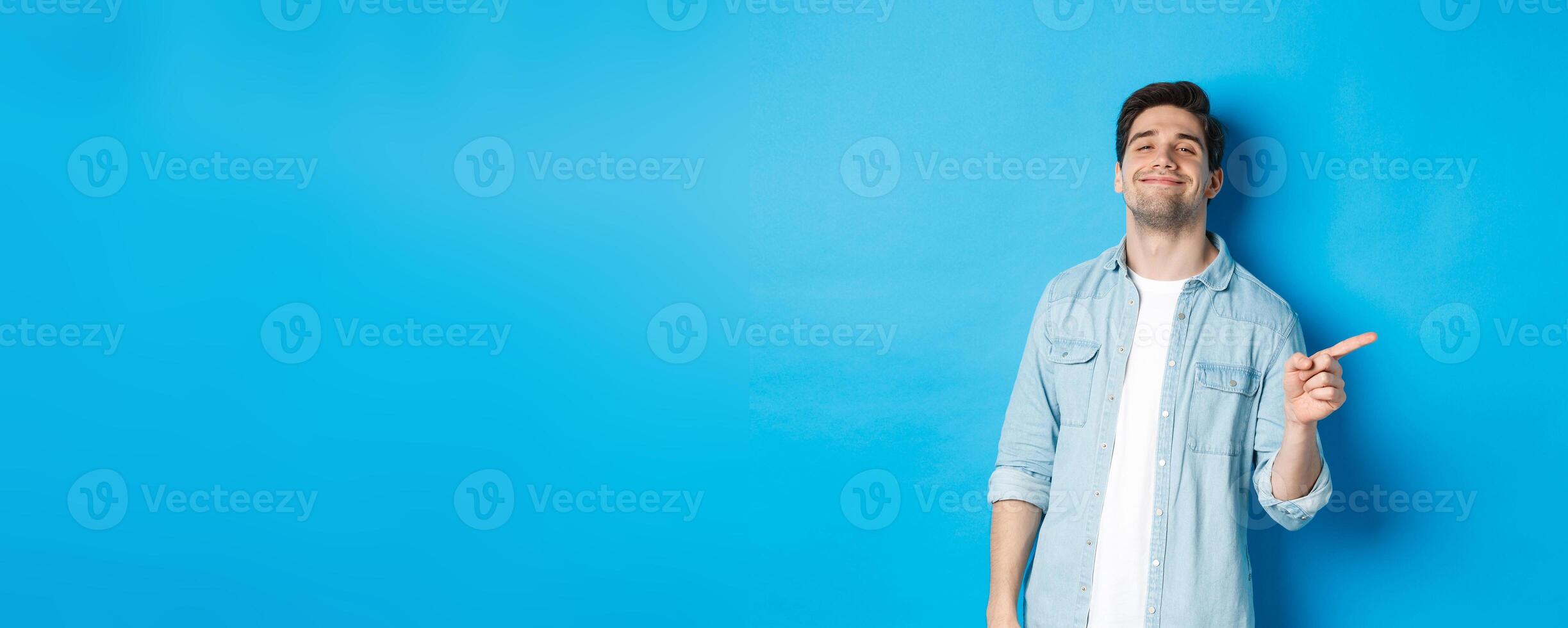 Confident handsome guy smiling sassy, pointing finger right at copy space, showing advertisement, standing over blue background photo