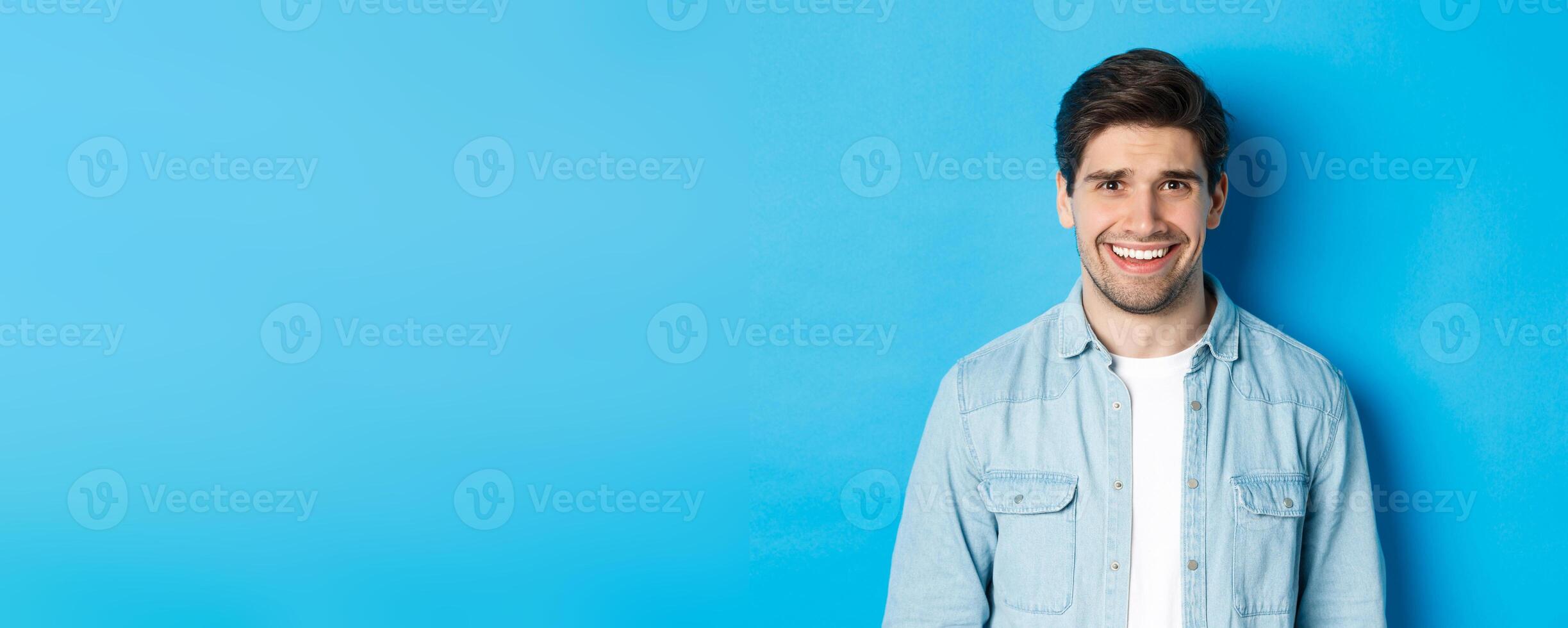 Close-up of young man feeling awkward, smile and cringe from uncomfortable situation, standing over blue background photo