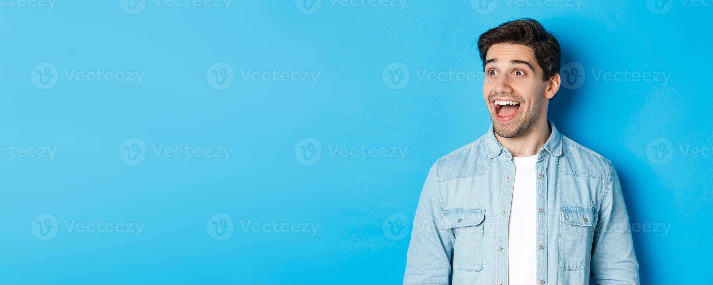 Close-up of handsome caucasian male model looking amazed left, checking out advertisement, standing over blue background photo