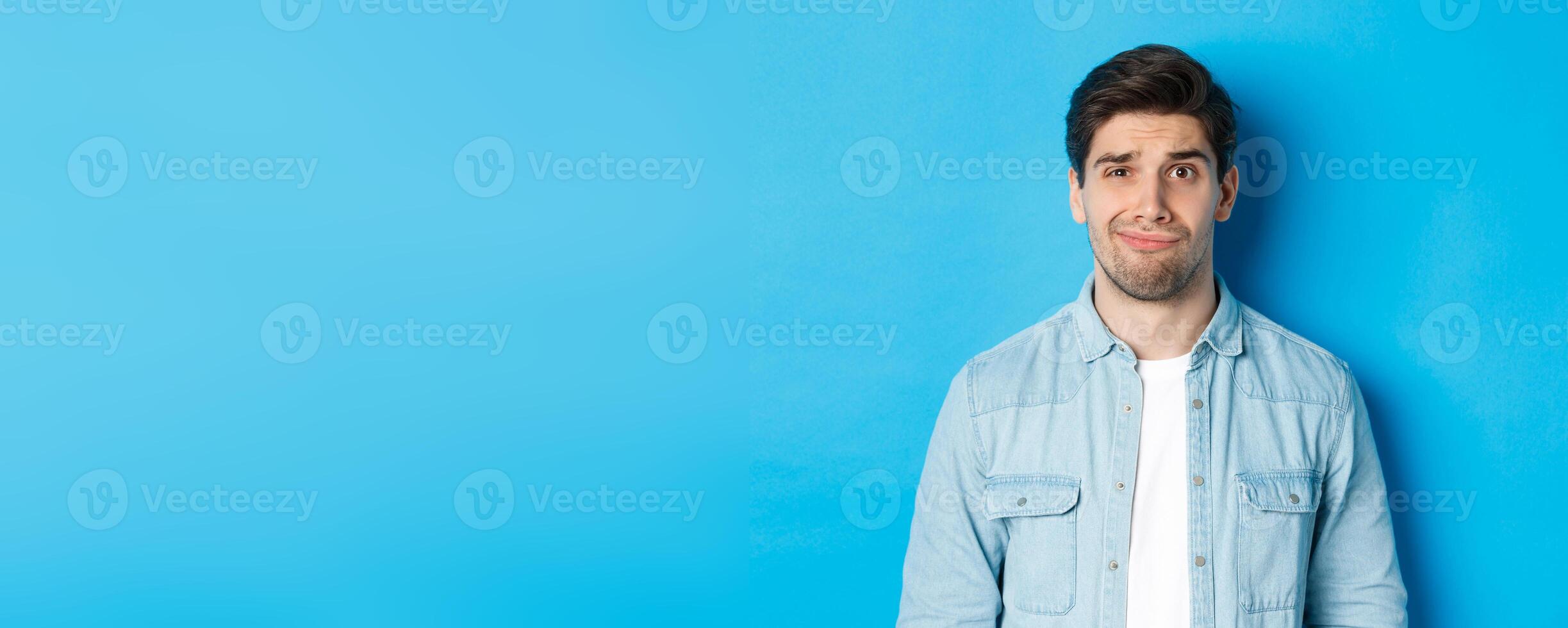 Close-up of skeptical and awkward guy smirking, feeling uncomfortable, standing over blue background photo