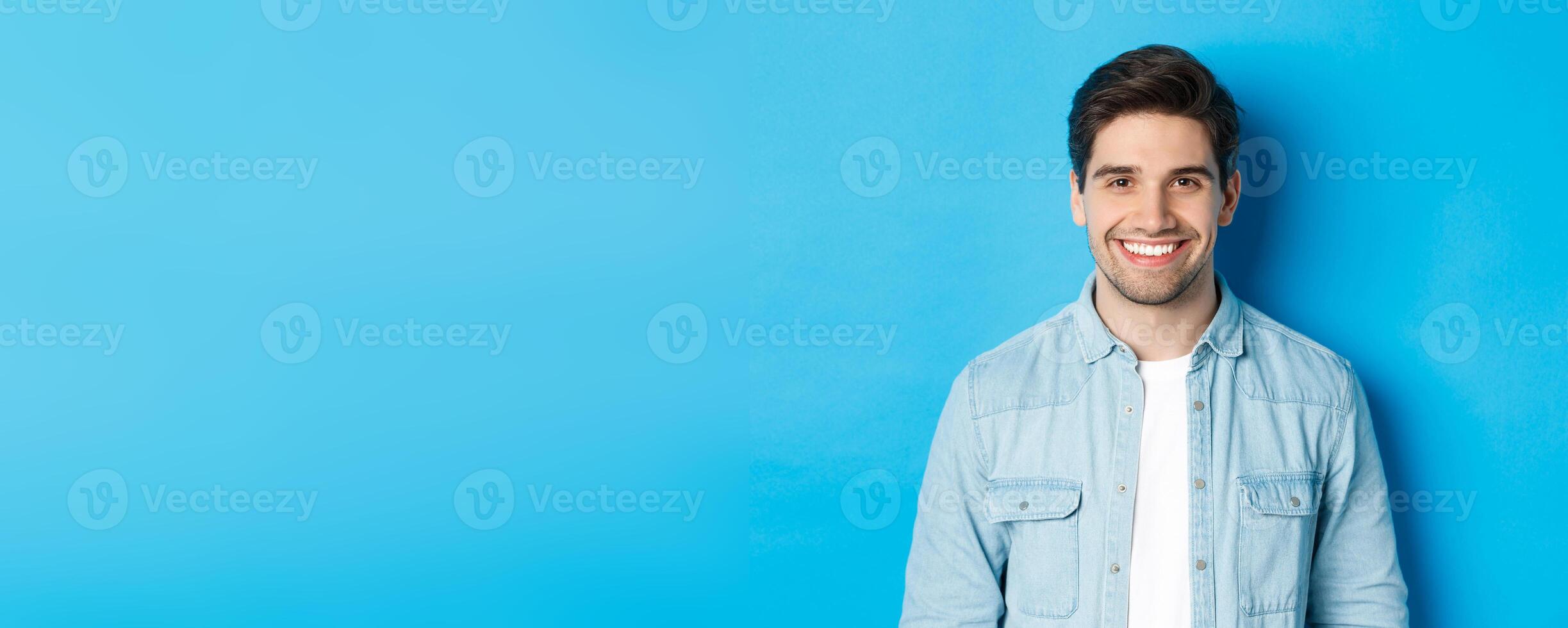Close-up of young successful man smiling at camera, standing in casual outfit against blue background photo