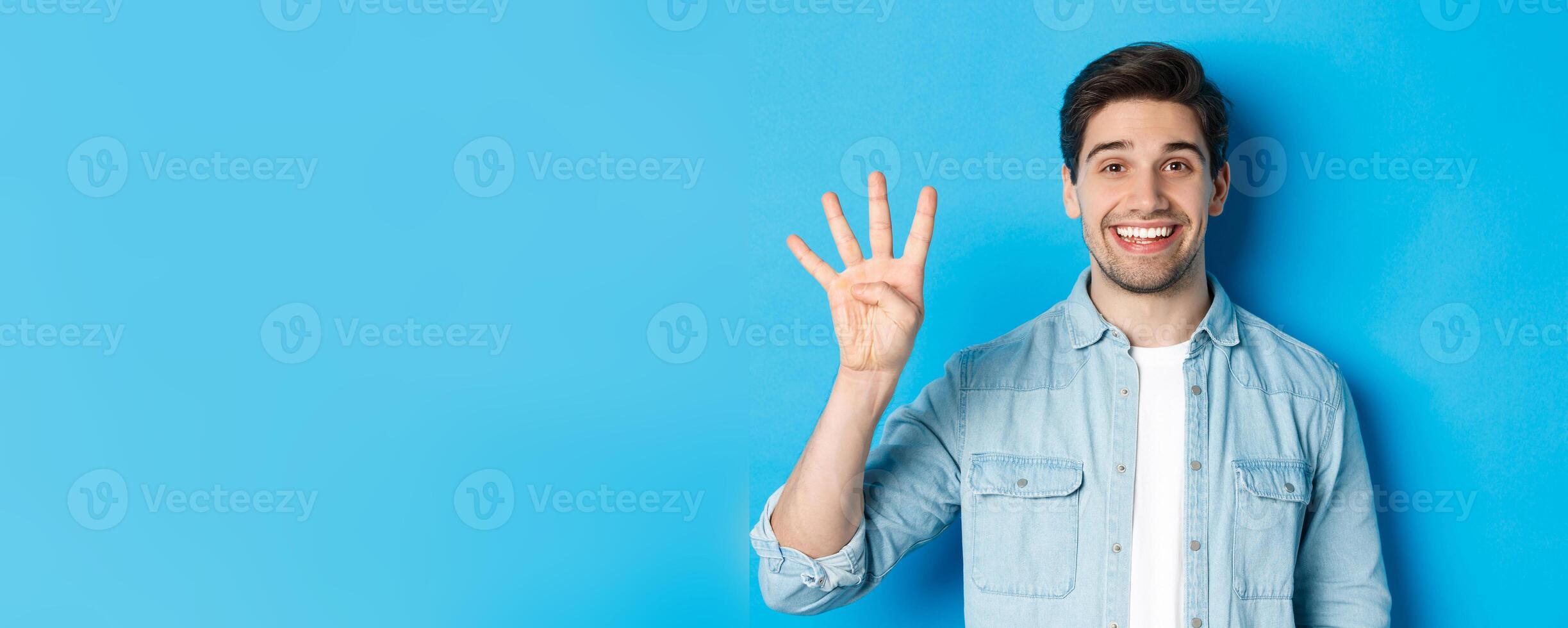 Close-up of handsome man smiling, showing fingers number four, standing over blue background photo
