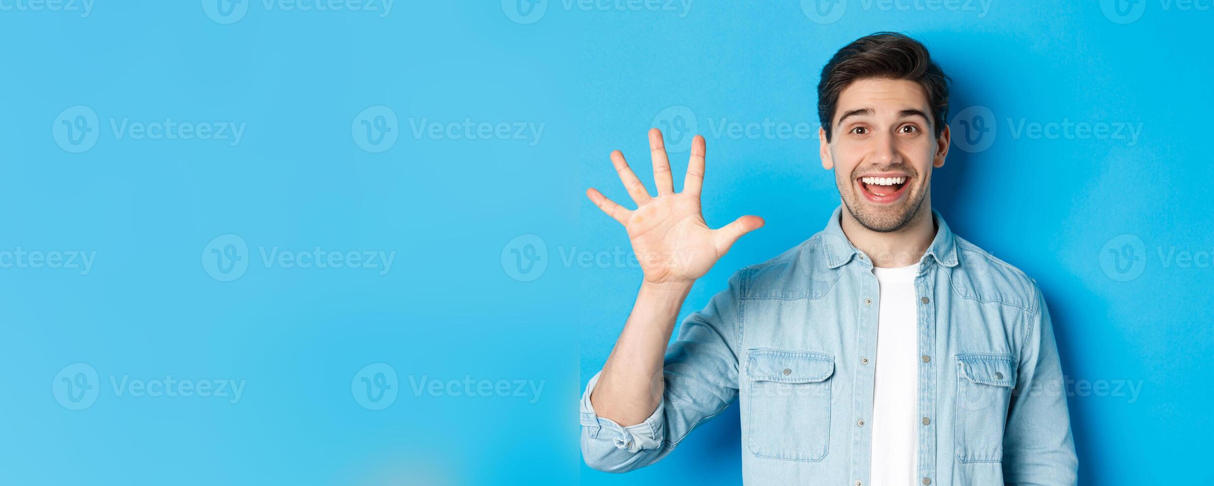 Close-up of handsome man smiling, showing fingers number five, standing over blue background photo