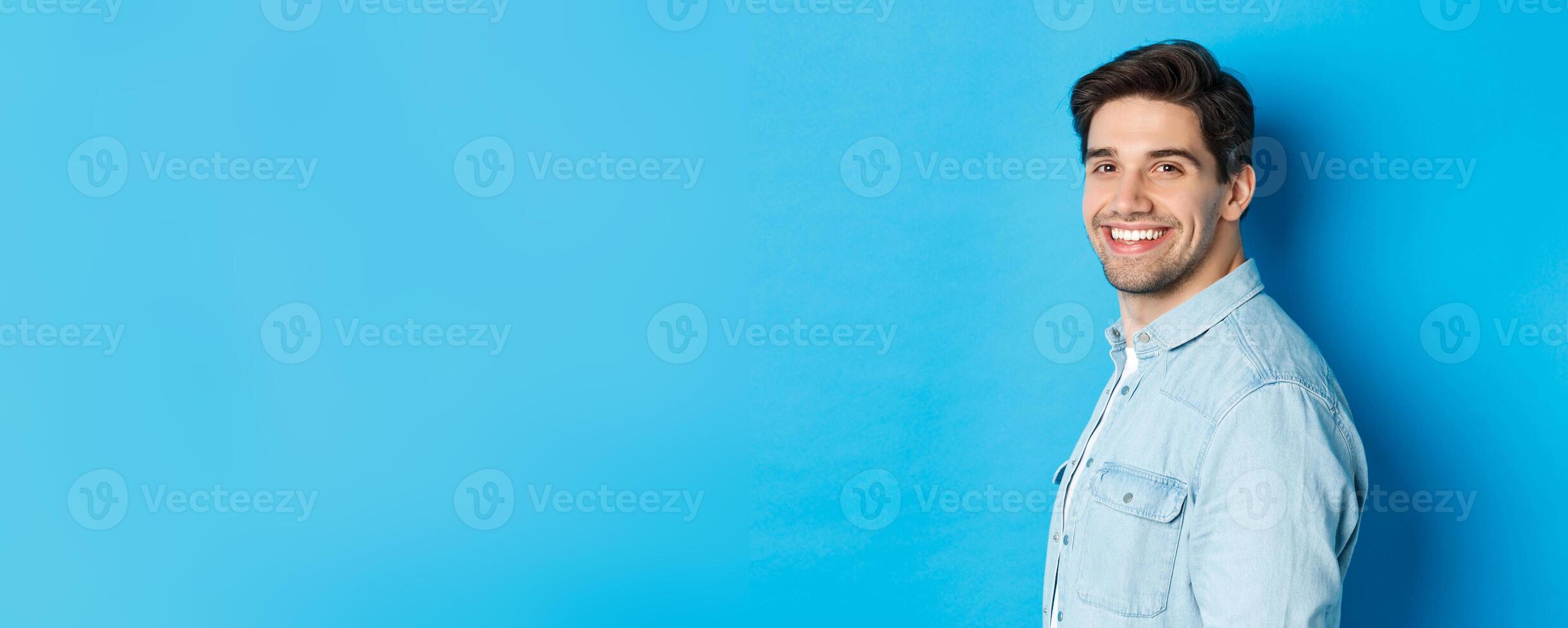 Handsome and confident guy turn head at camera, smiling happy, standing over blue background photo