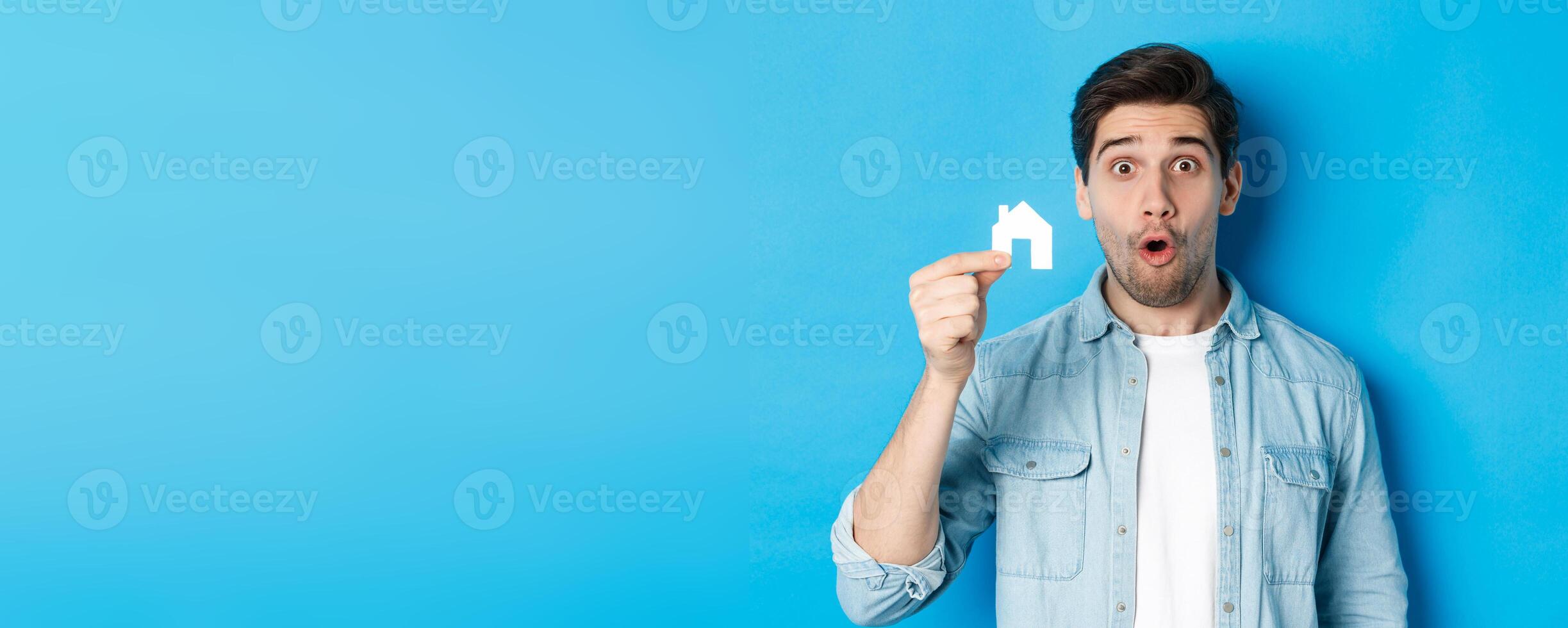 Real estate concept. Surprised guy showing small paper house model, looking amazed, standing against blue background photo