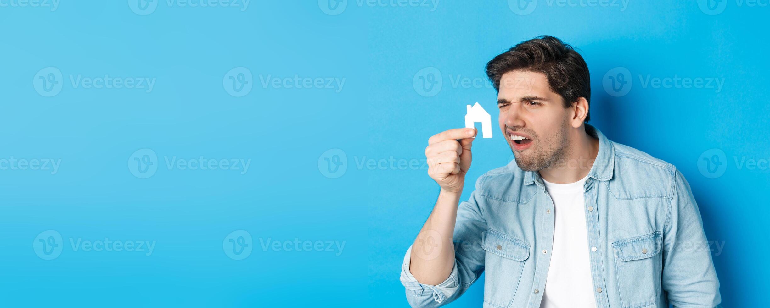 Real estate concept. Man looking at tiny house maket with confused face, standing puzzled against blue background photo
