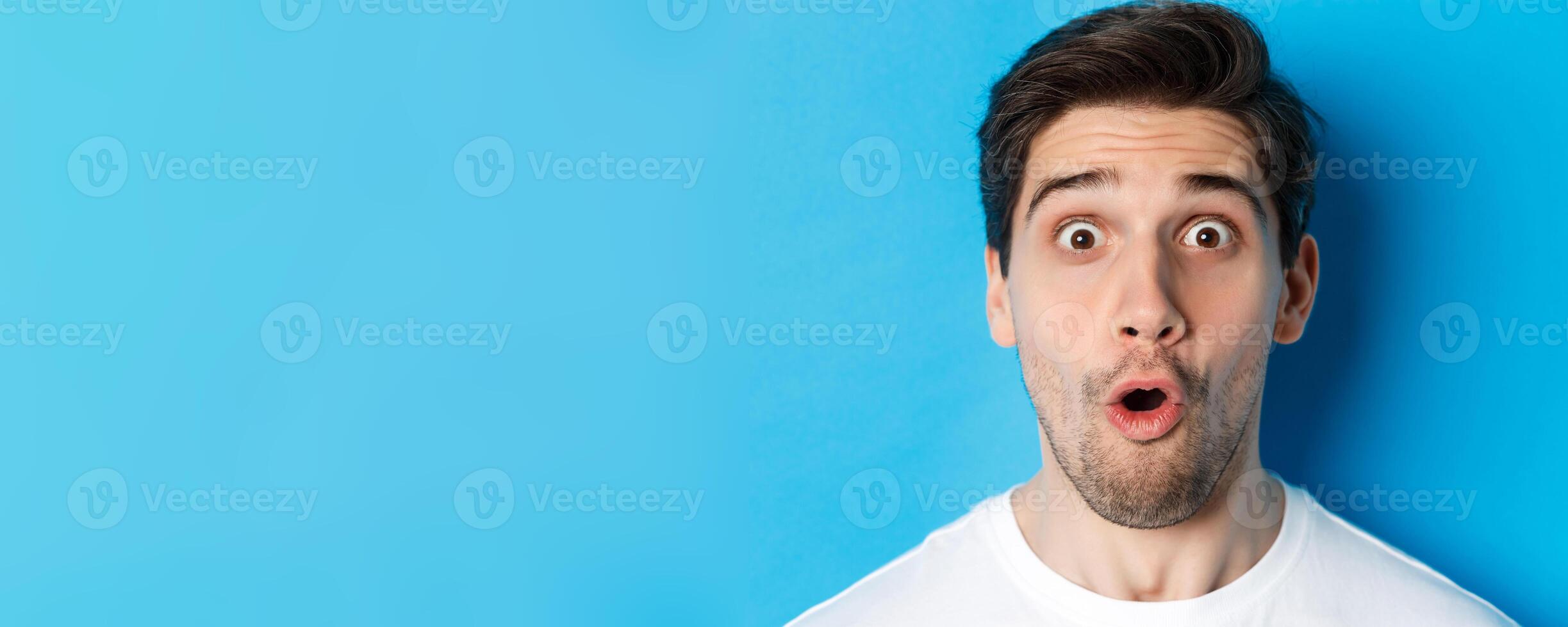Headshot of surprised man gasping wondered, saying wow and looking amazed at camera, standing over blue background photo