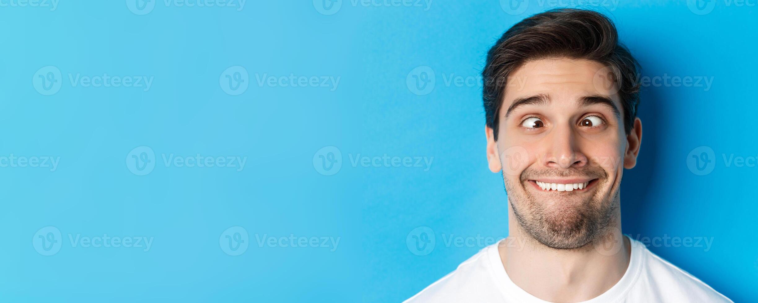 Head shot of young man making funny expressions, smiling and squinting, standing over blue background photo