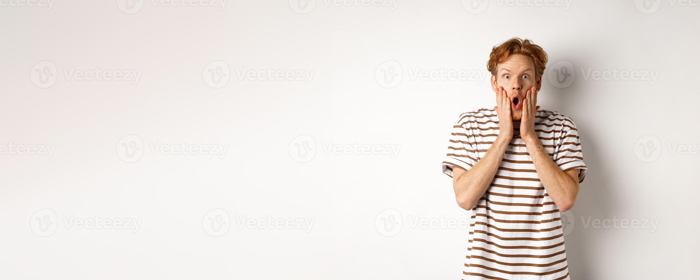 Impressed young man with ginger hair, gasping and staring shocked at camera, express complete disbelief and amazement, white background photo