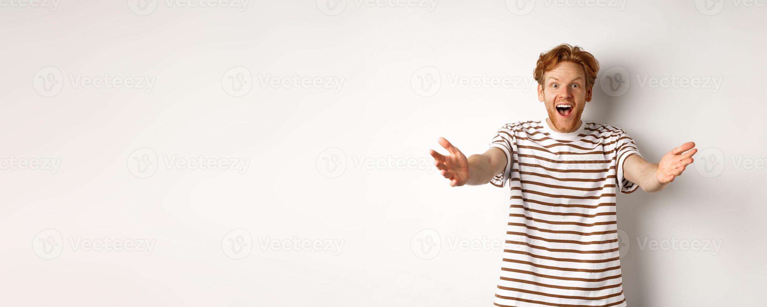 Cheerful man with red curly hair reaching hands forward, stretch out arms to welcome or congratulate you, smiling happy, standing over white background photo