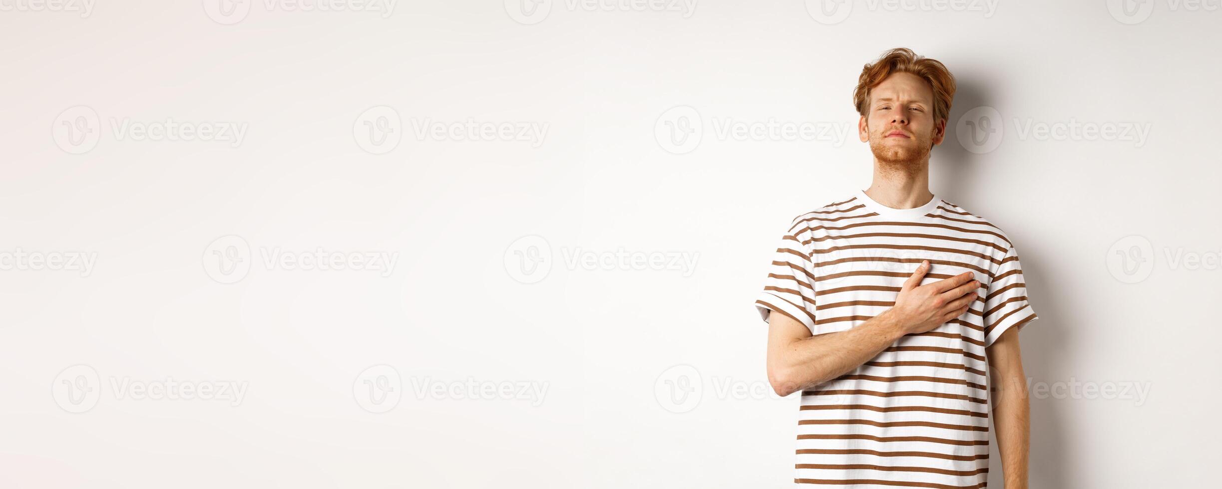 Young bearded redhead man holding hand on heart and looking patriotic, standing during national anthem, standing over white background photo