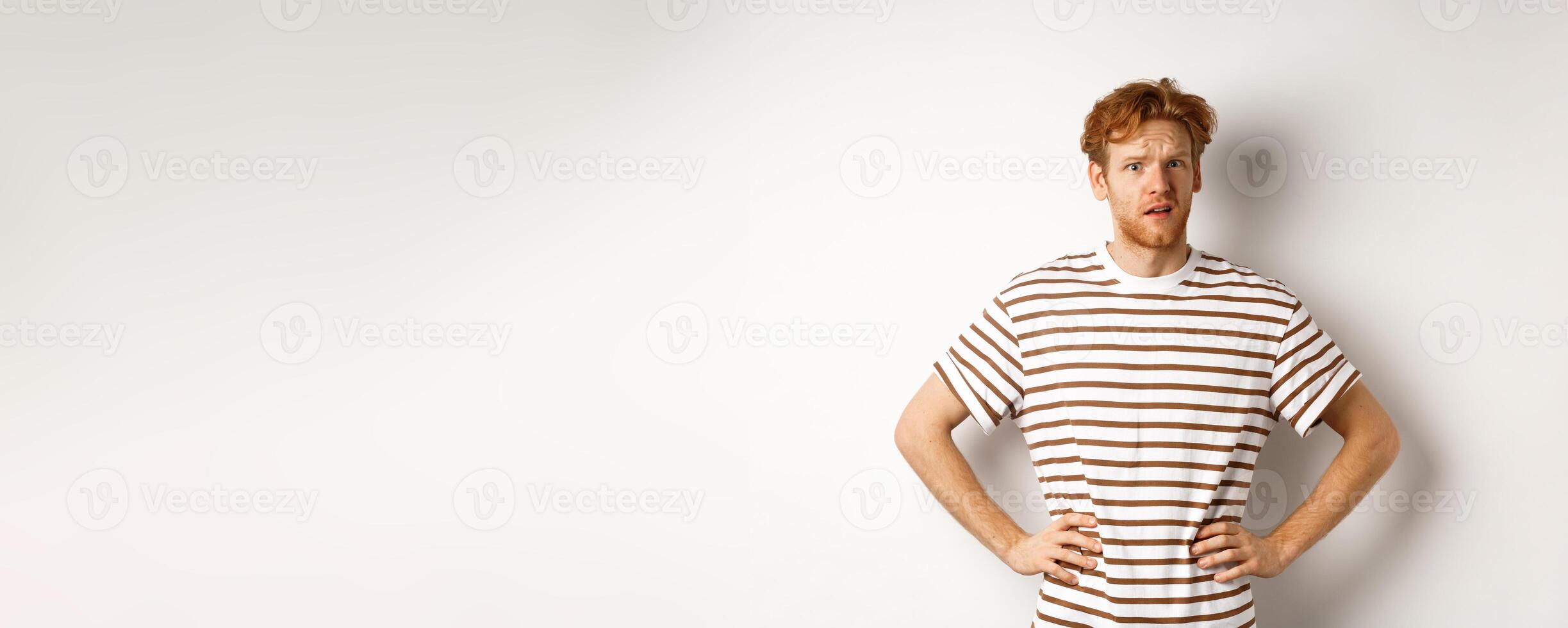 Confused and offended man with ginger curly hair staring at camera puzzled, standing questioned against white background photo