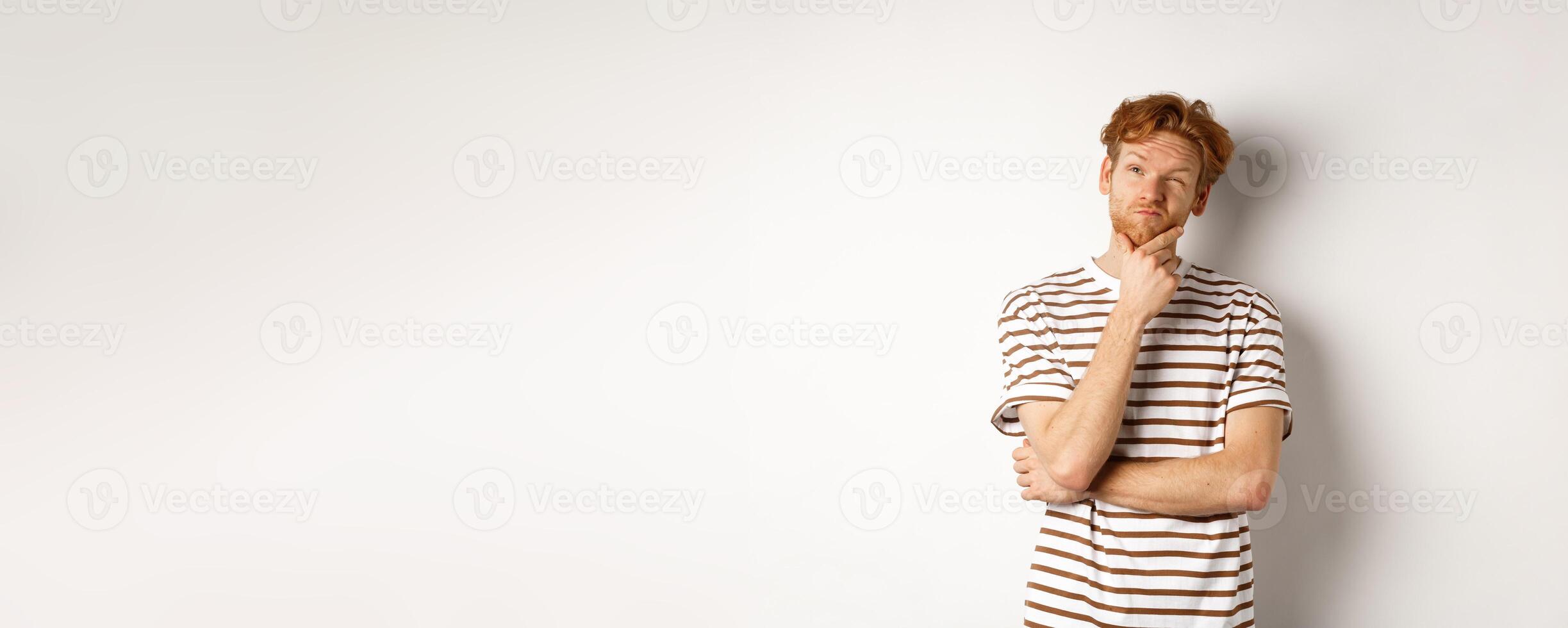 Portrait of thoughtful man with red hair thinking, looking at upper left corner and making choice, standing over white background photo