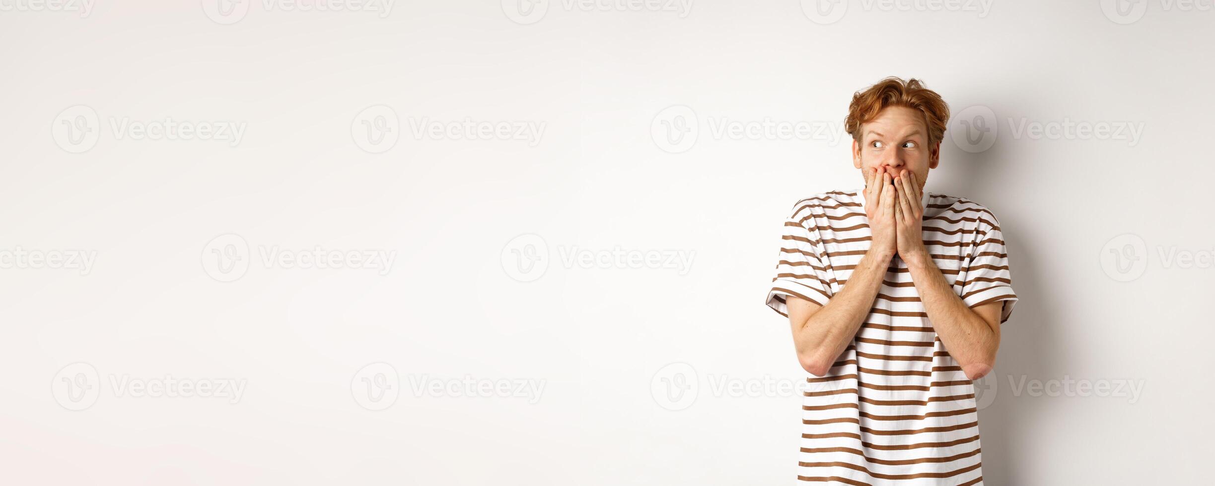 Shocked redhead guy gossiping, giggle in hands and looking left impressed, standing over white background photo