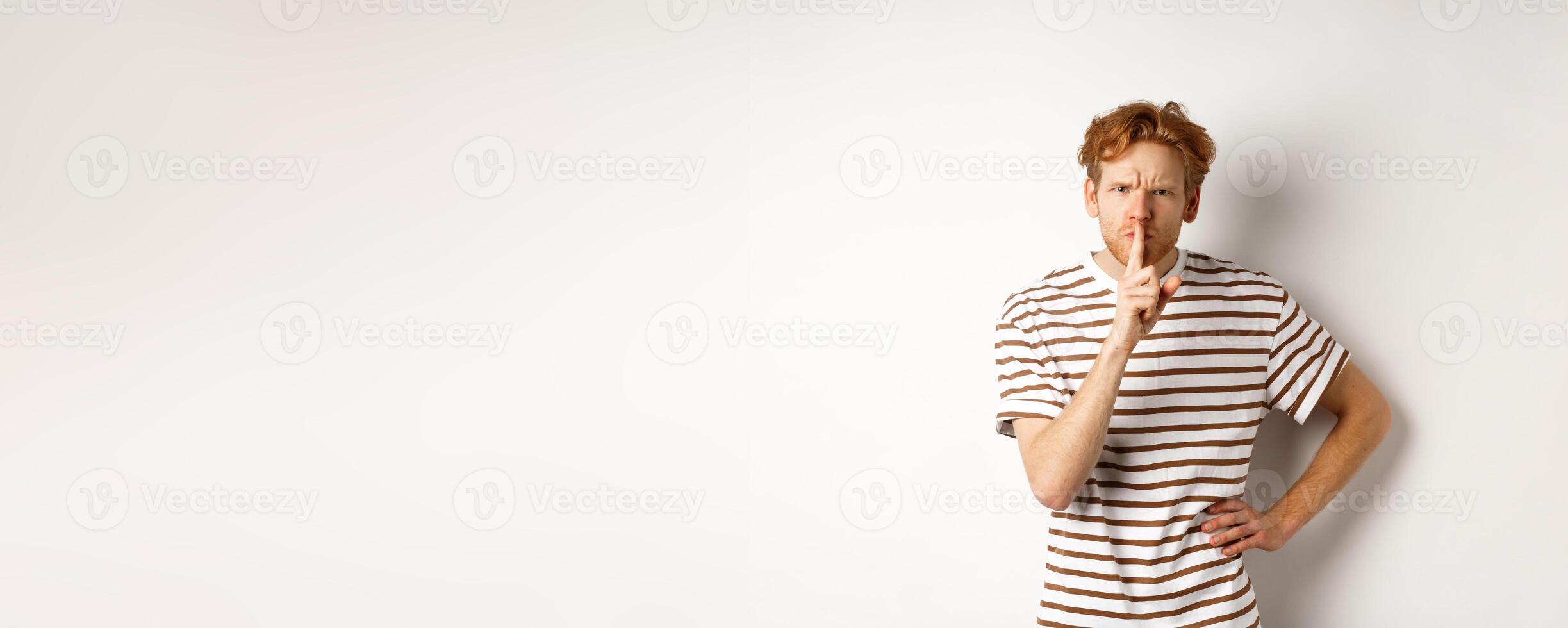 Angry redhead man looking displeased and hushing at camera, making shh gesture, standing over white background photo