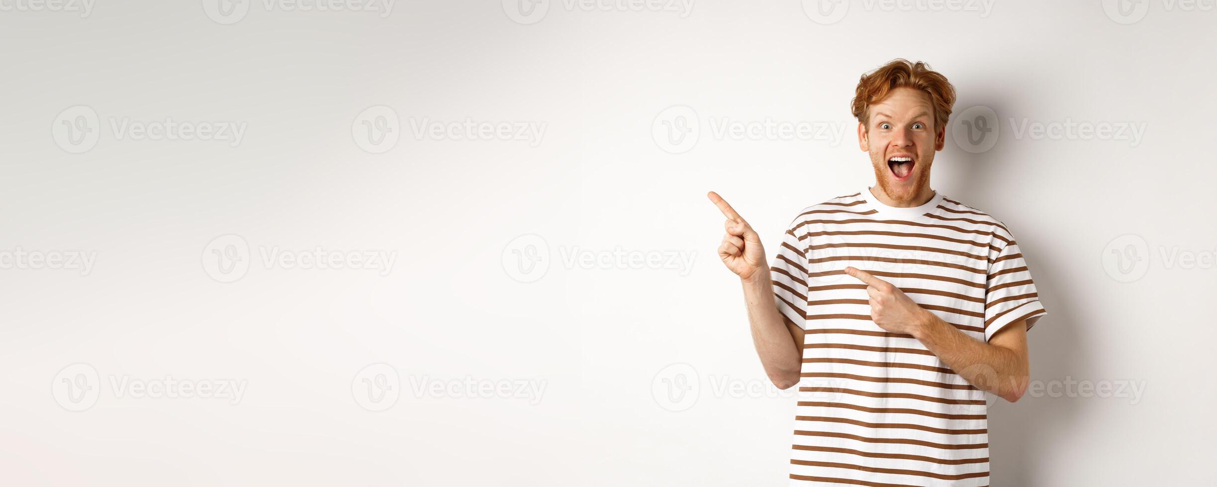 Cheerful man with red hair checking out promo offfer, pointing left at logo and smiling at camera, standing over white background photo