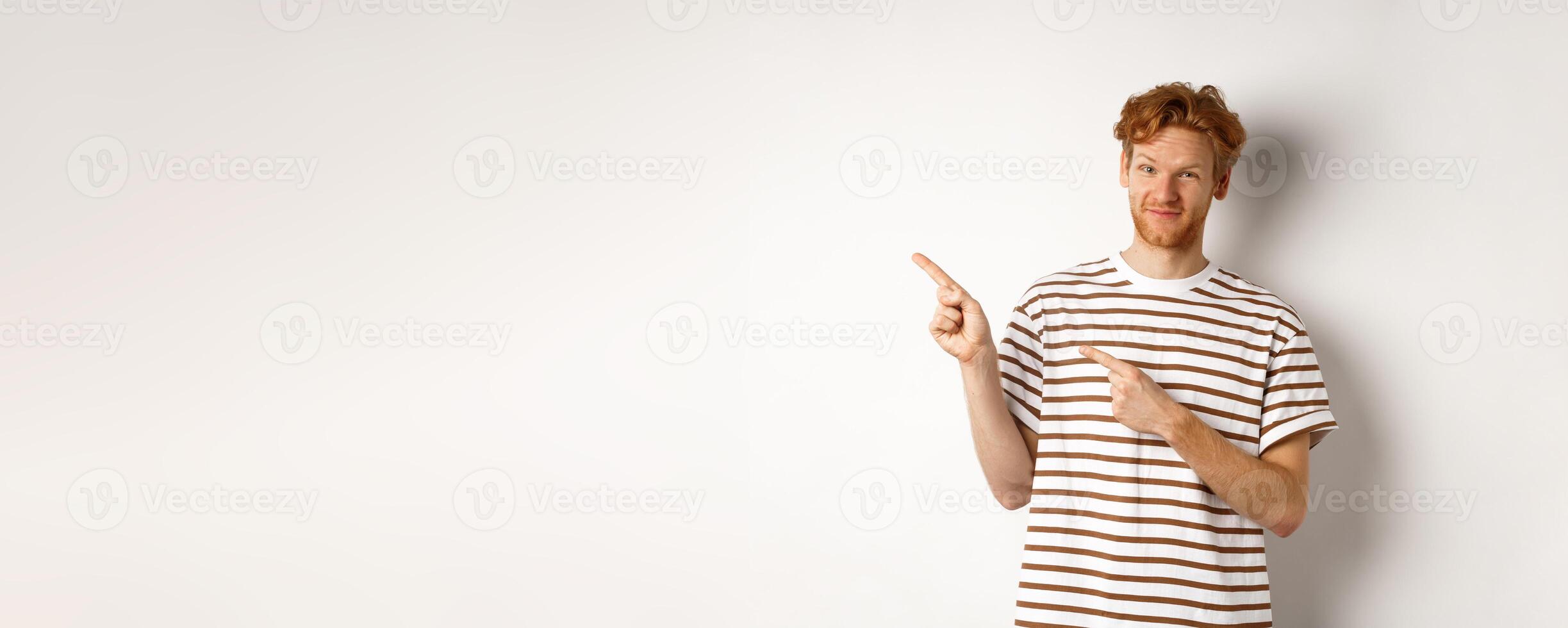 Doubtful redhead guy pointing fingers left at looking skeptical at camera, standing over white background photo