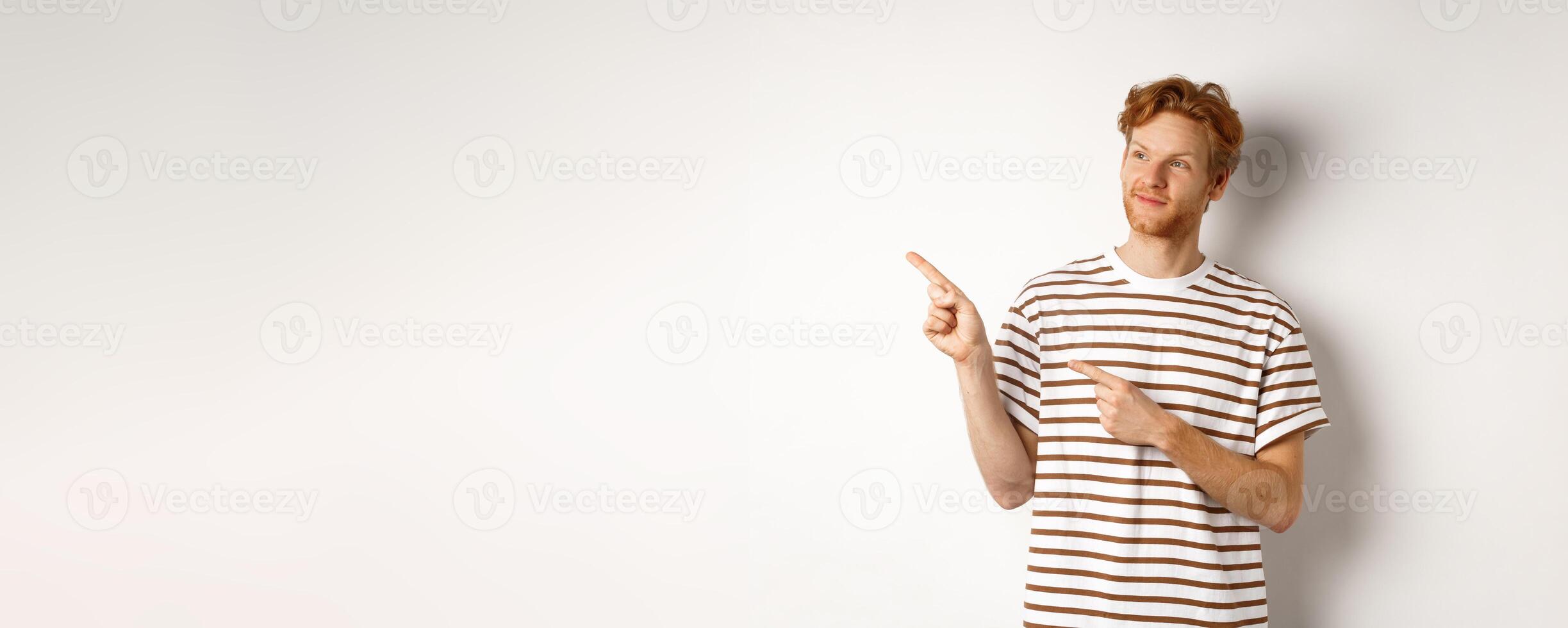 Smiling man with curly red hair, wearing striped t-shirt, smiling and pointing fingers left, demonstrate banner, standing over white background photo