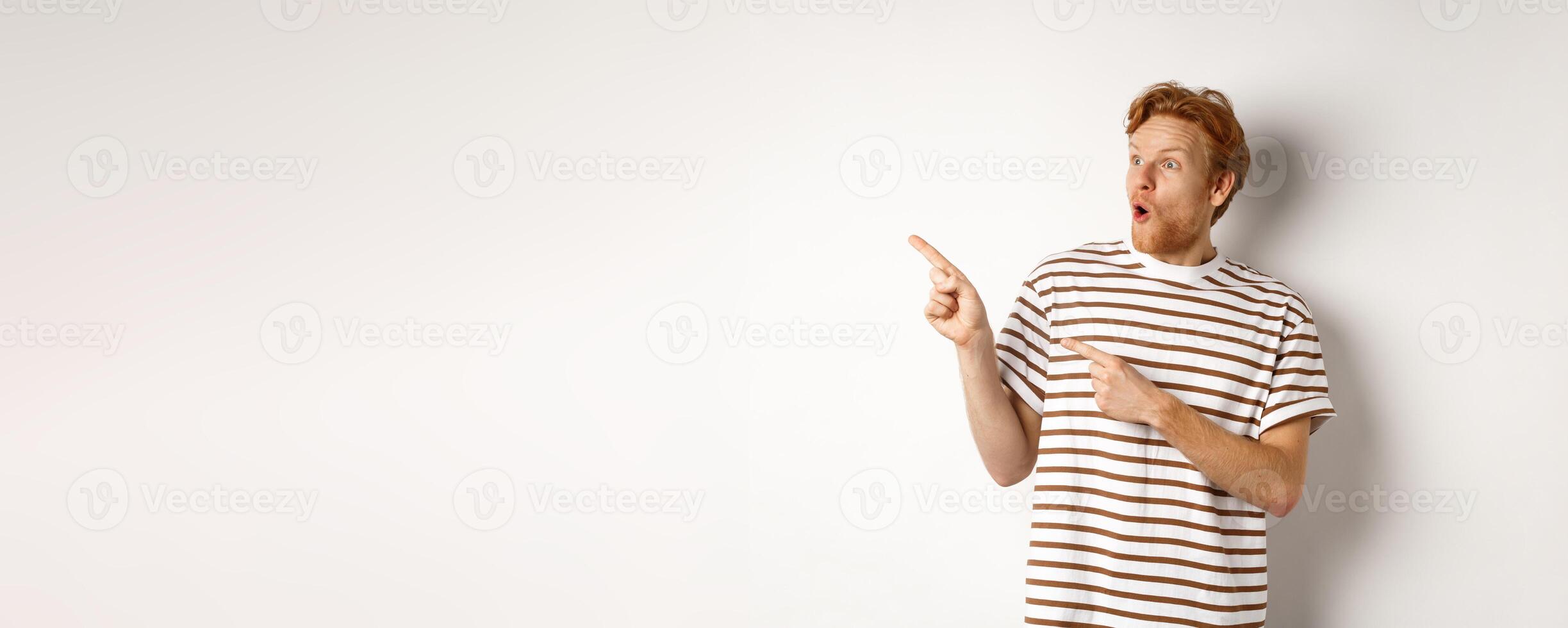 Check it out. Amazed redhead guy pointing and looking left at promotion banner, saying wow, standing over white background photo