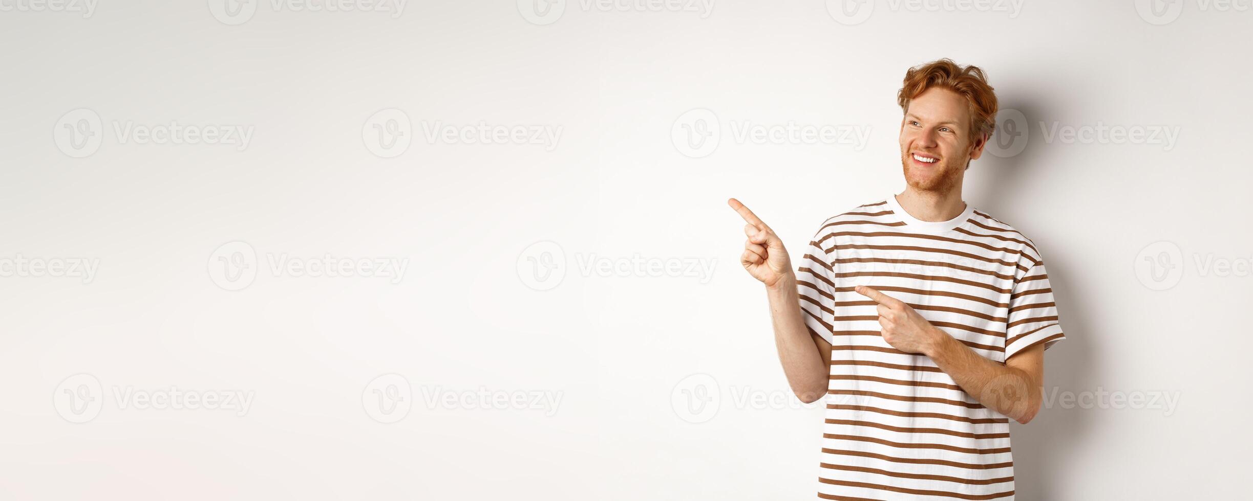 Cheerful caucasian guy with red hair and beard, looking and pointing left at logo banner, standing over white background photo