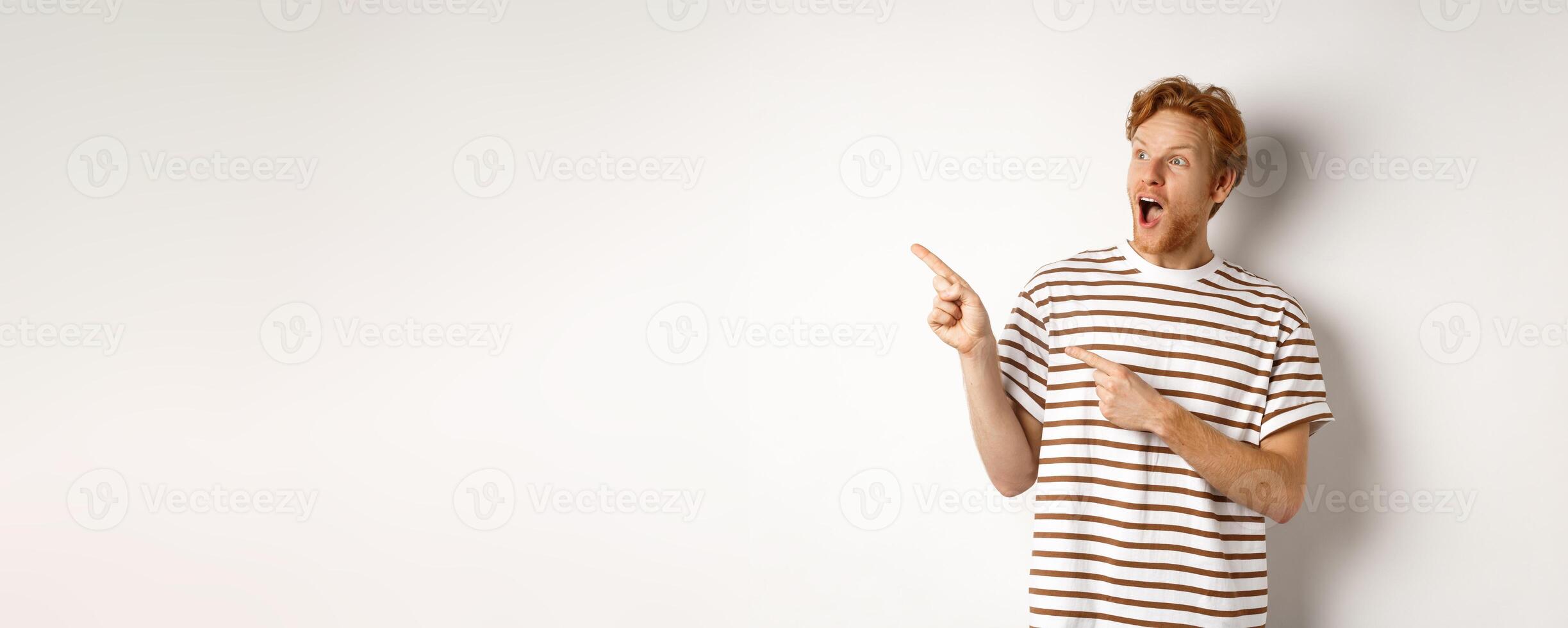 Check it out. Amazed redhead guy pointing and looking left at promotion banner, standing over white background photo