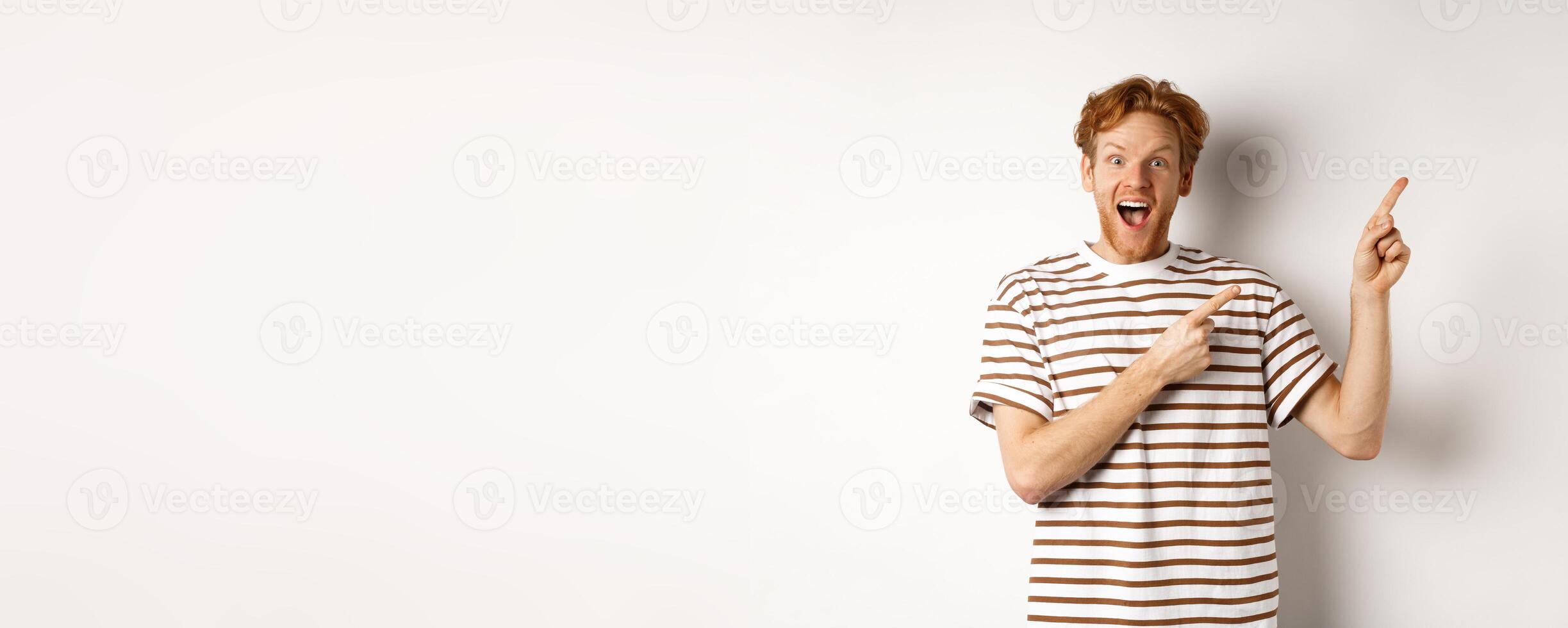 Excited redhead guy in striped t-shirt checking out special deal, pointing fingers right and smiling at camera, white background photo