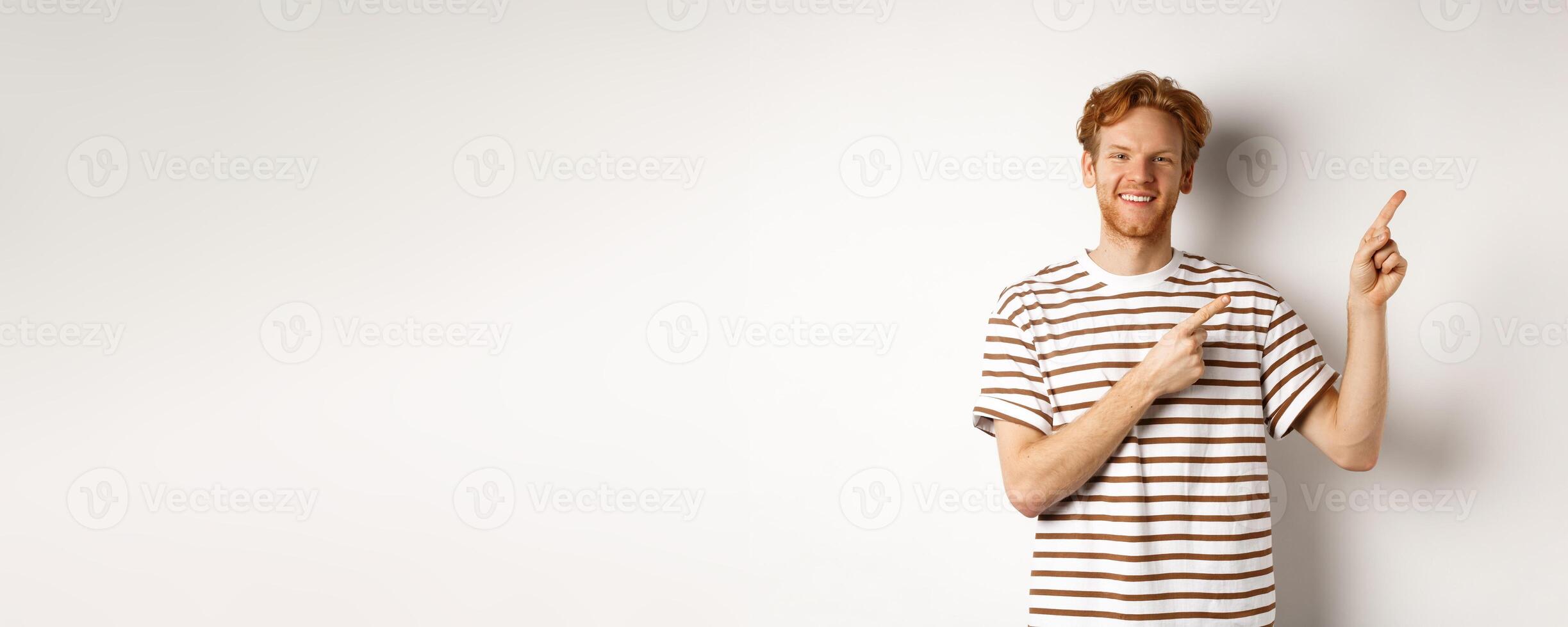 Handsome redhead man showing logo, pointing fingers right and smiling, standing over white background photo