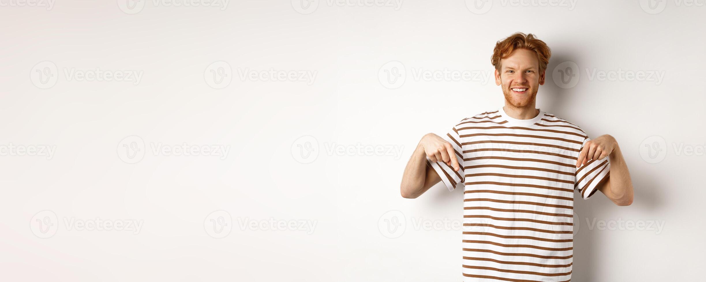 Smiling young man with messy red hair and beard, pointing fingers down, recommending shop, standing over white background photo