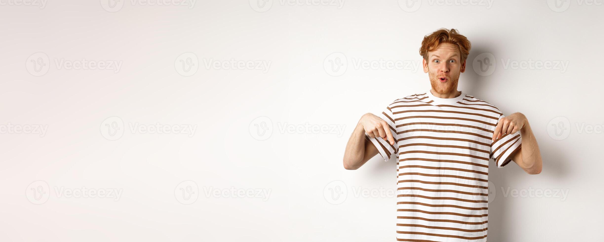 Image of funny redhead male student pointing fingers down, showing promo offer with excited smile, standing over white background photo