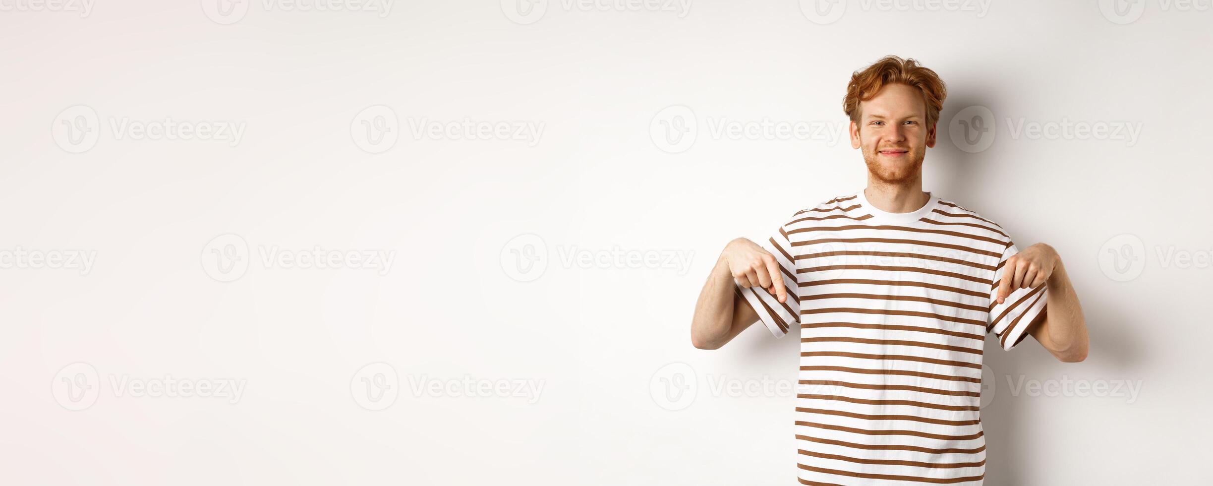 Handsome male model with ginger hair and bristle, pointing fingers down and smiling, showing advertisement, white background photo
