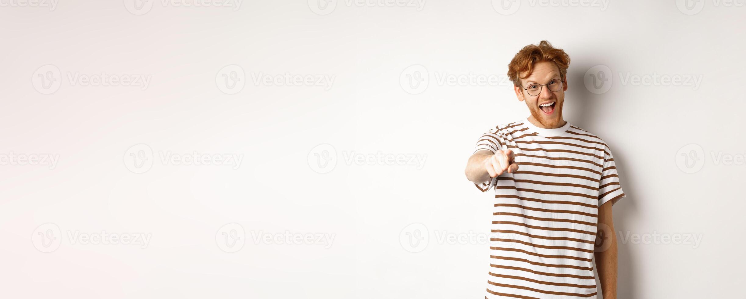 Image of funny redhead guy in glasses laughing at you, pointing finger camera and making fun of someone, standing over white background photo
