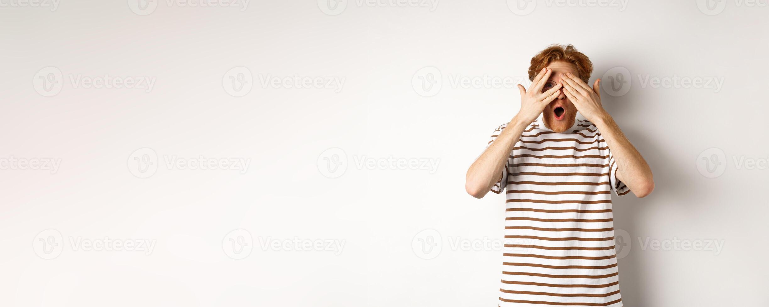 Shocked and embarrassed redhead guy covering eyes, peeking through fingers and gasping, standing over white background photo