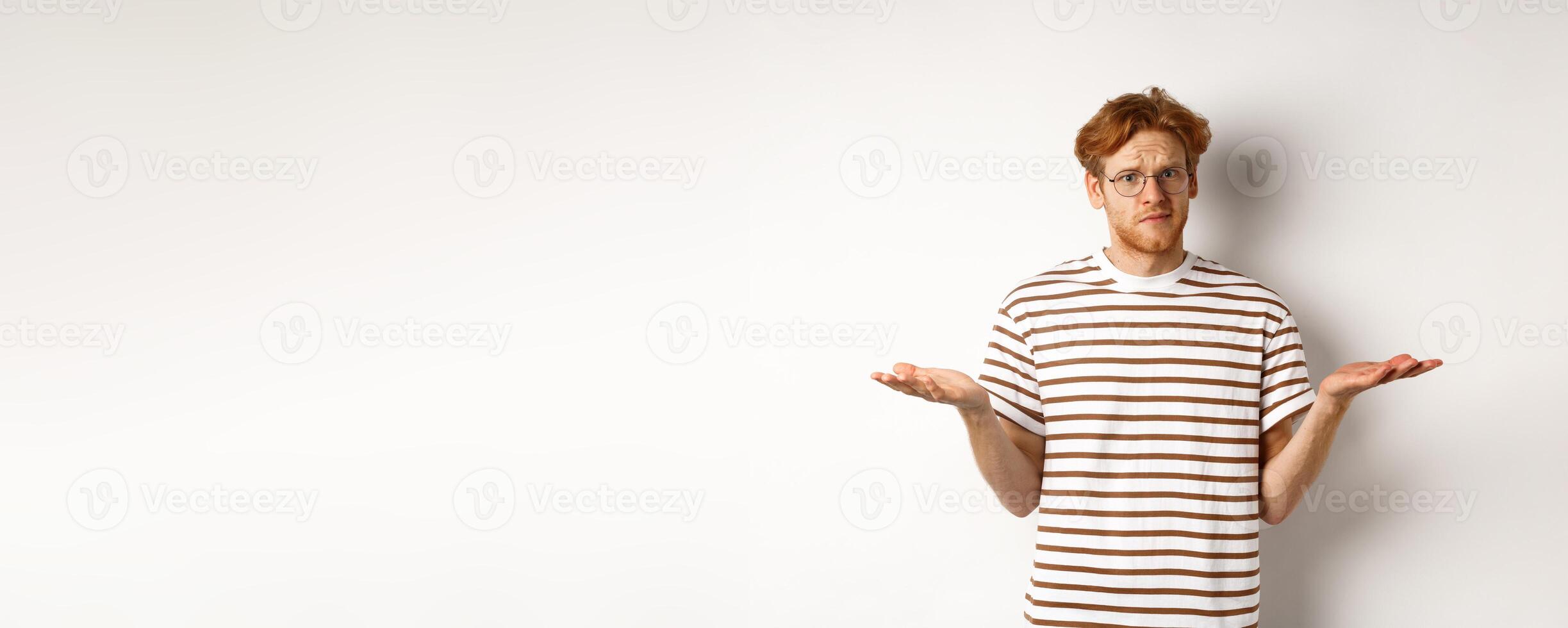 Confused boyfriend with red hair shrugging, spread hands sideways and staring at camera puzzled, asking what, stnading over white background photo